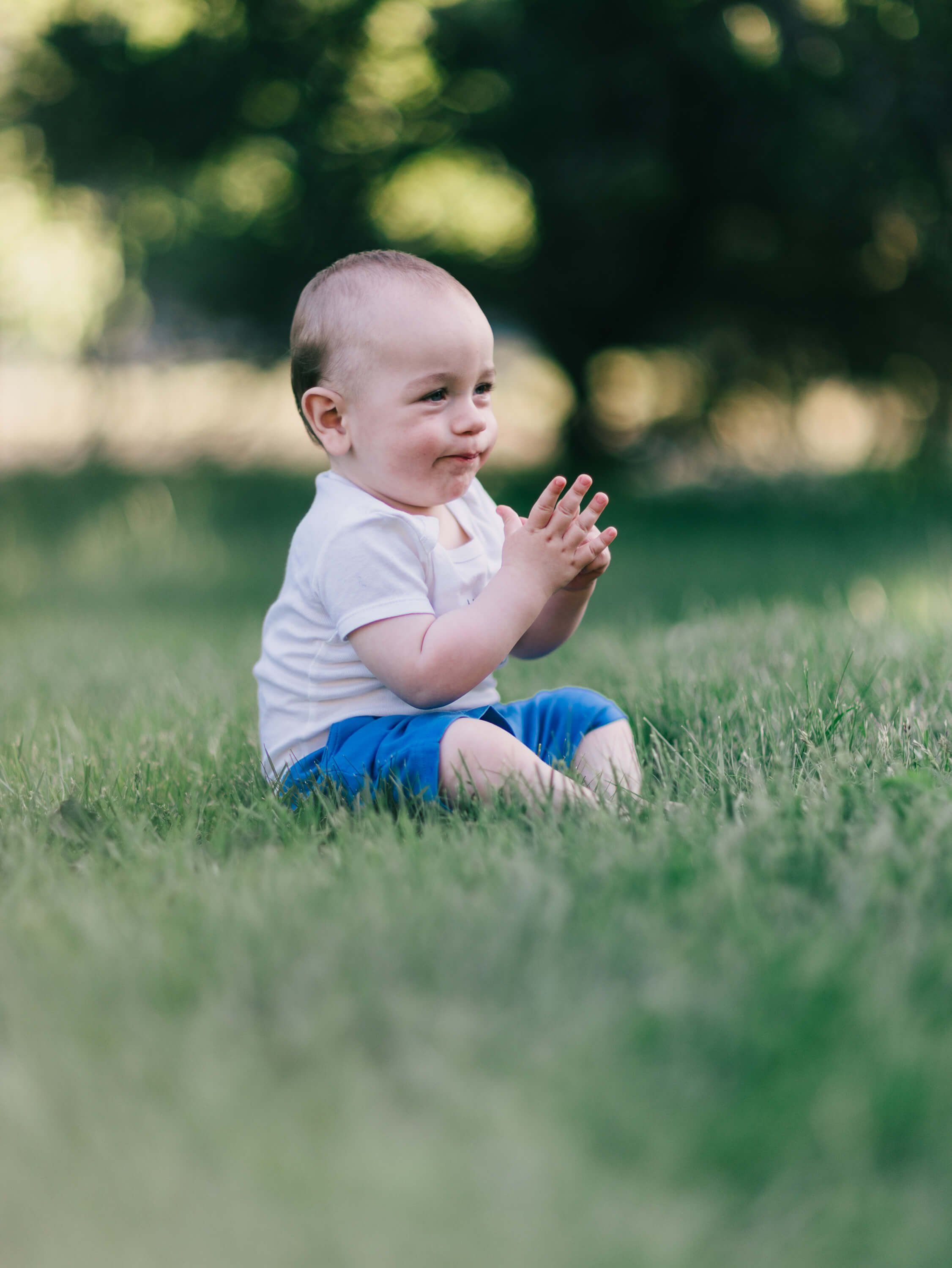 Smiling toddler in white shirt and blue pants sits on grass with soft, dappled light filtering through trees behind