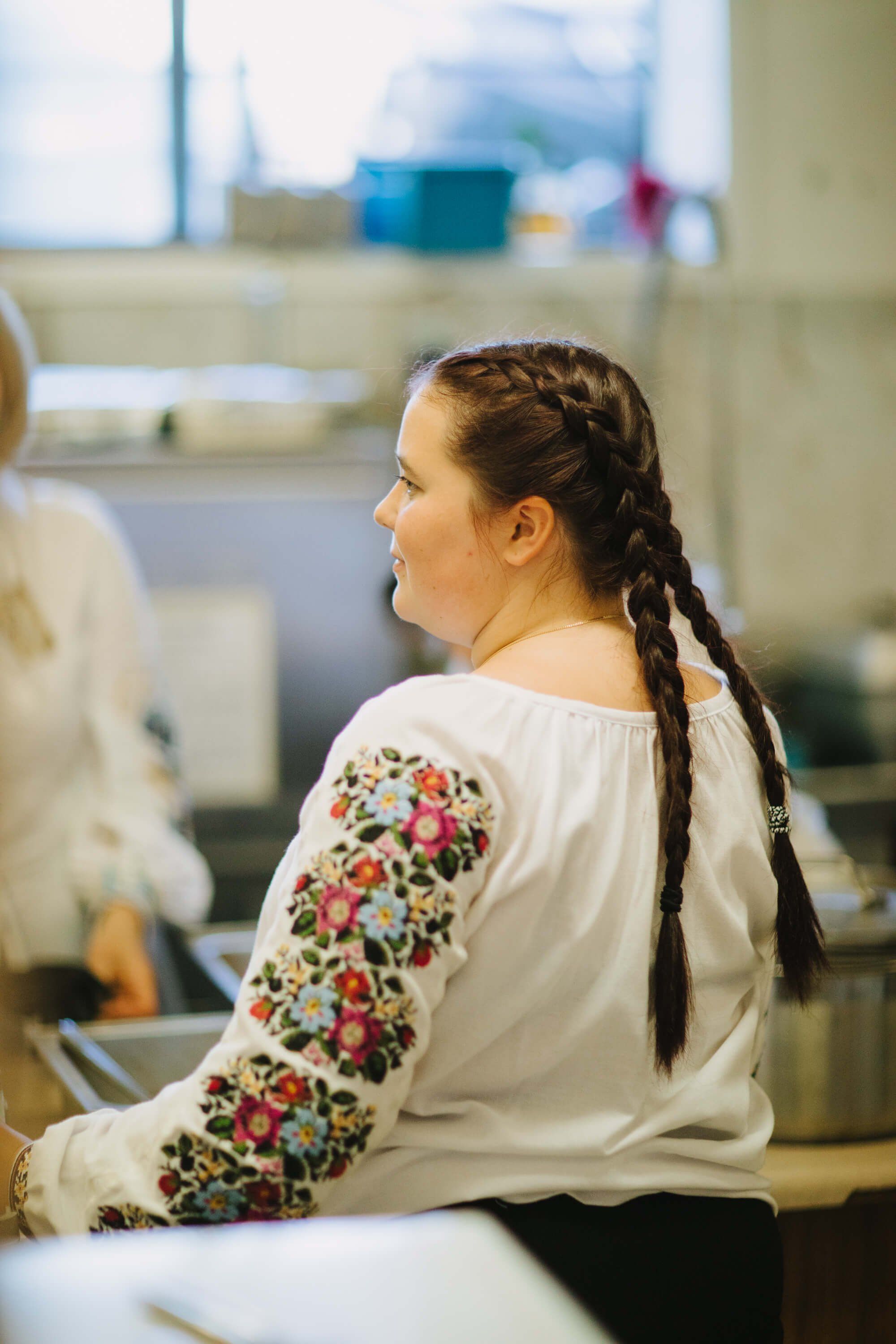 Woman in white embroidered blouse with braided hair sits at kitchen counter during Ukrainian fundraiser dinner