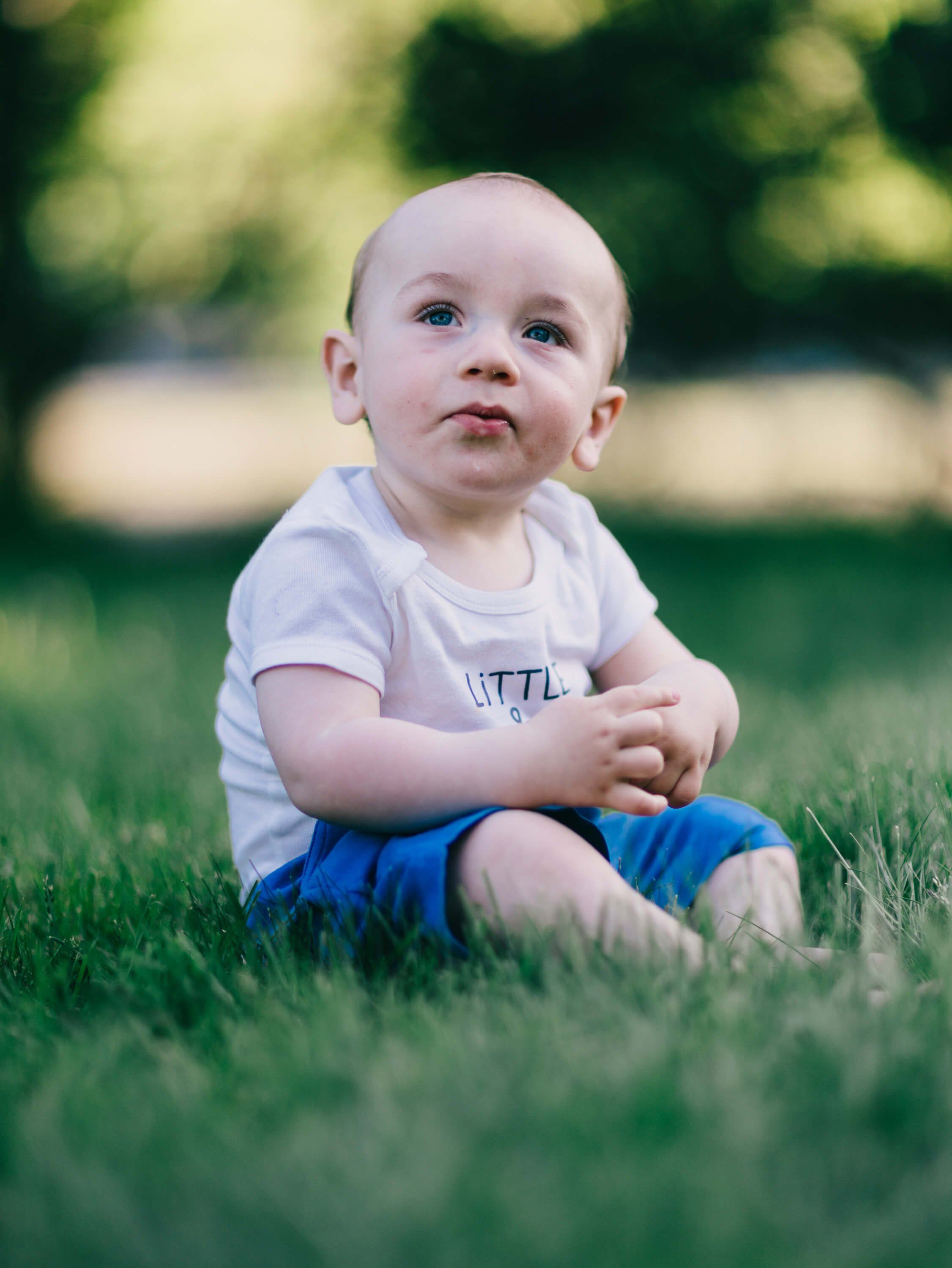 Toddler in white shirt and blue shorts sits on grass, gazing upward with soft natural light and blurred green background