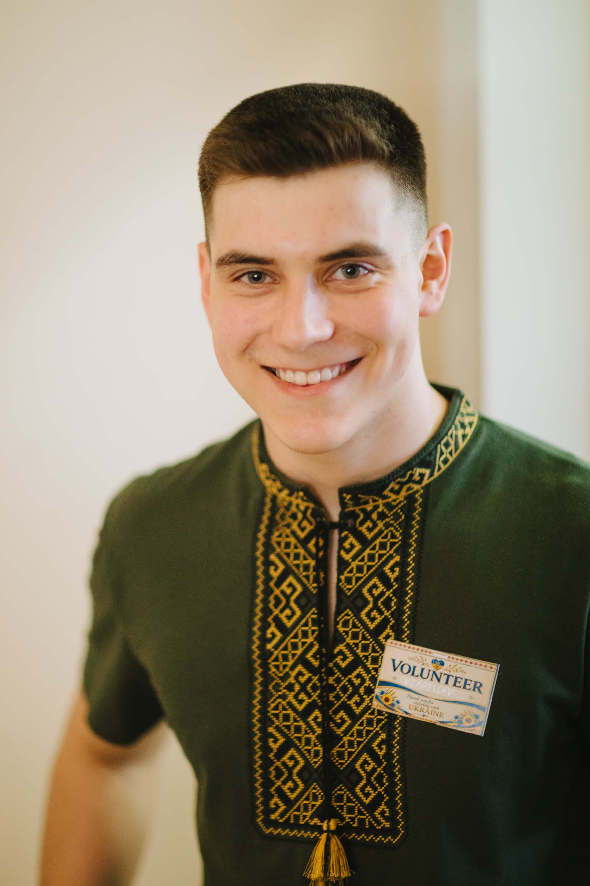 Smiling volunteer in olive green embroidered shirt with name badge stands against neutral background at fundraiser event