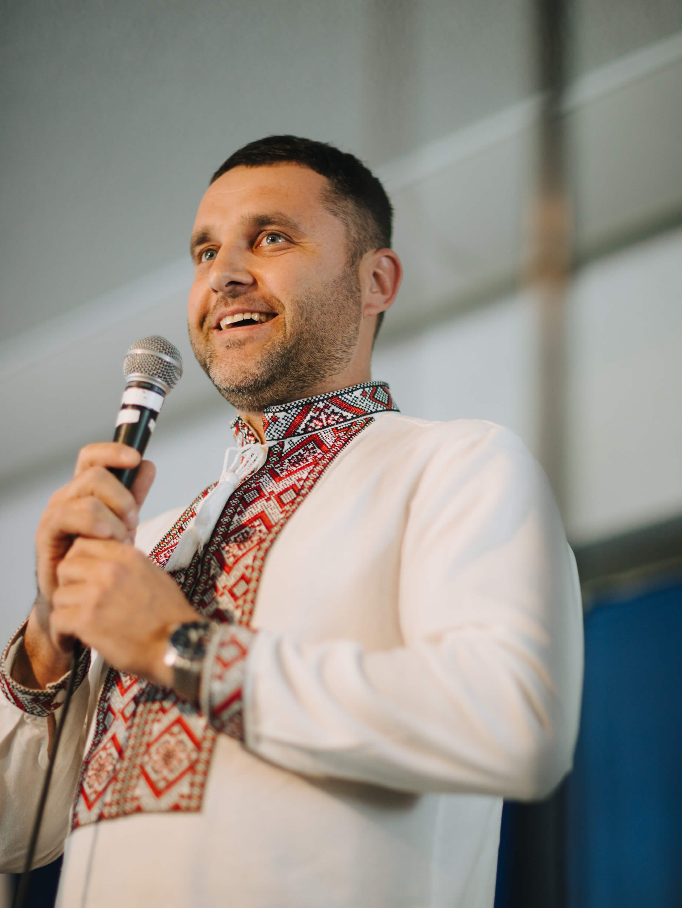 Man in embroidered Ukrainian shirt speaking into microphone at indoor fundraiser event
