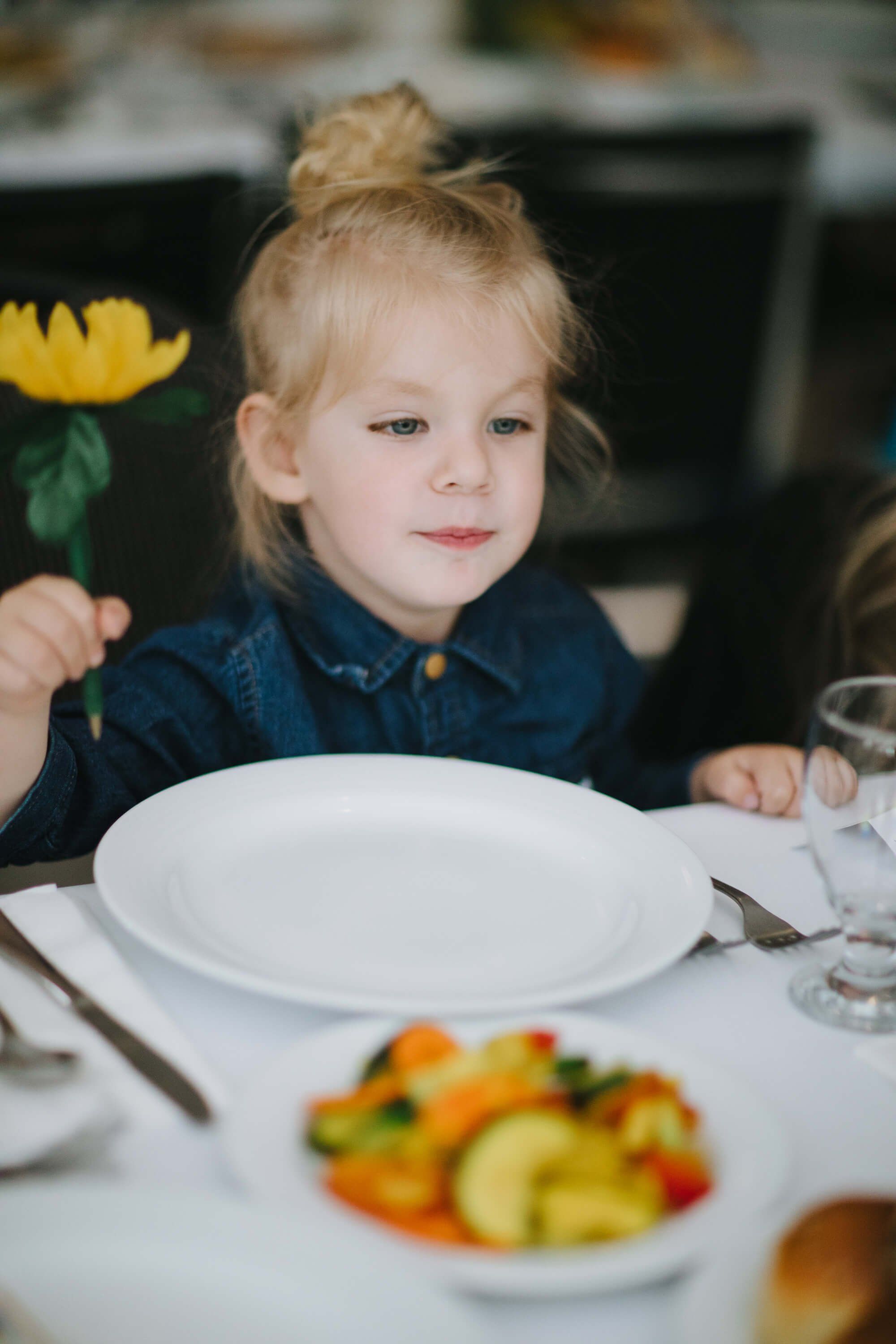 Young child with blonde hair in a bun at a formal dinner table, holding a yellow flower, with an empty plate and vegetables nearby