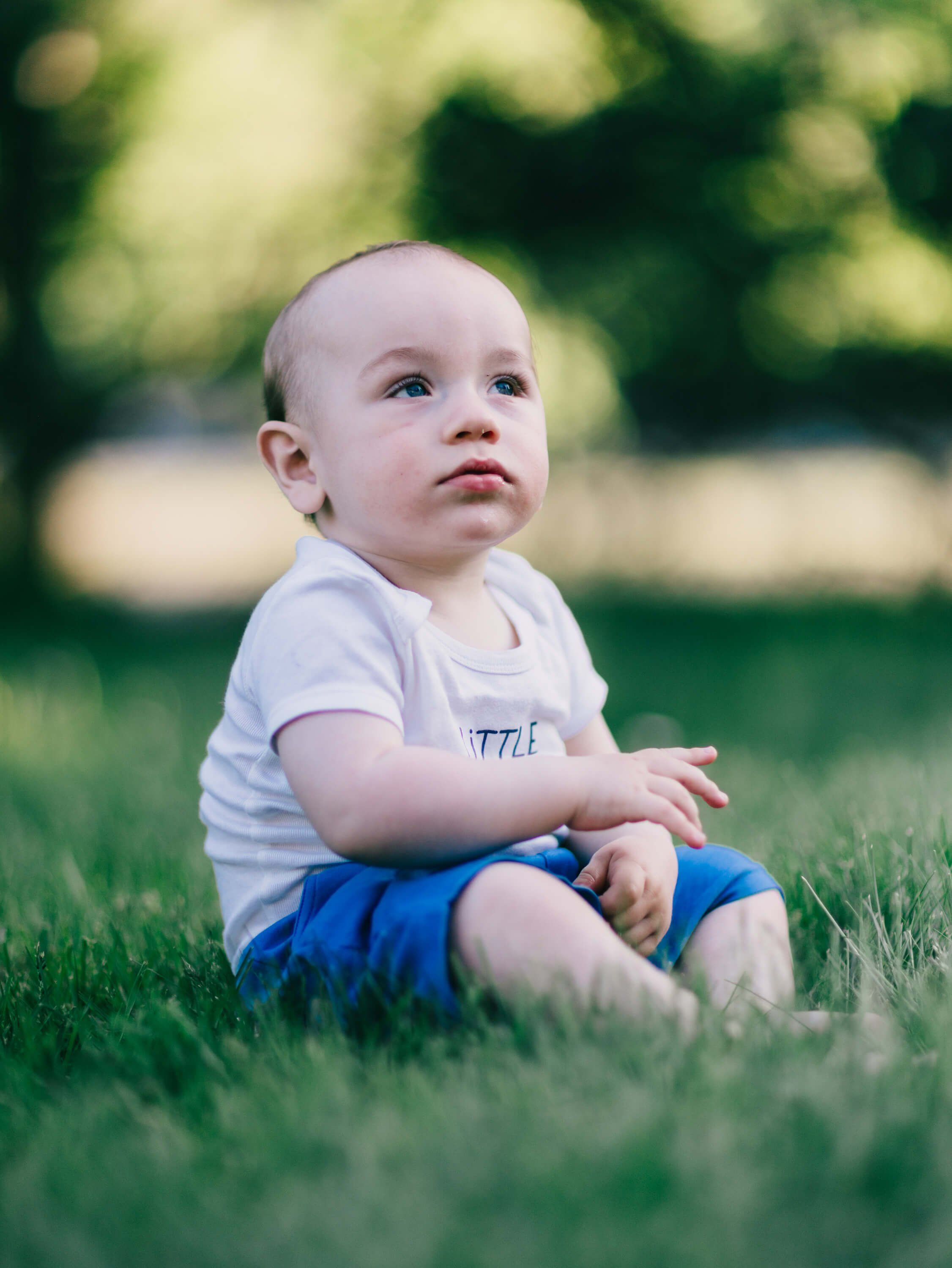 Toddler in white shirt and blue shorts sits on grass, gazing upward with soft natural light and blurred green foliage behind