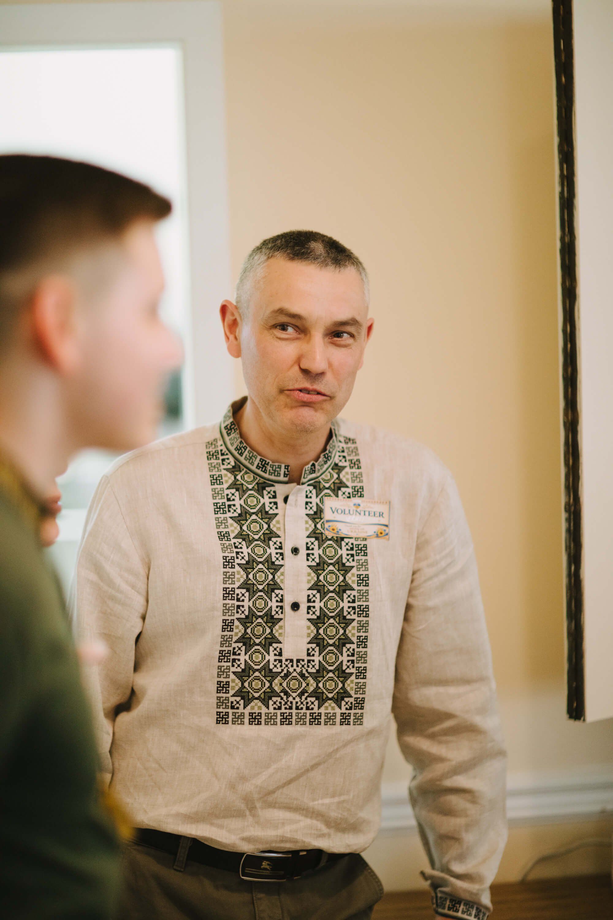 Man in embroidered linen shirt listens intently during conversation at indoor evening event, warm lighting