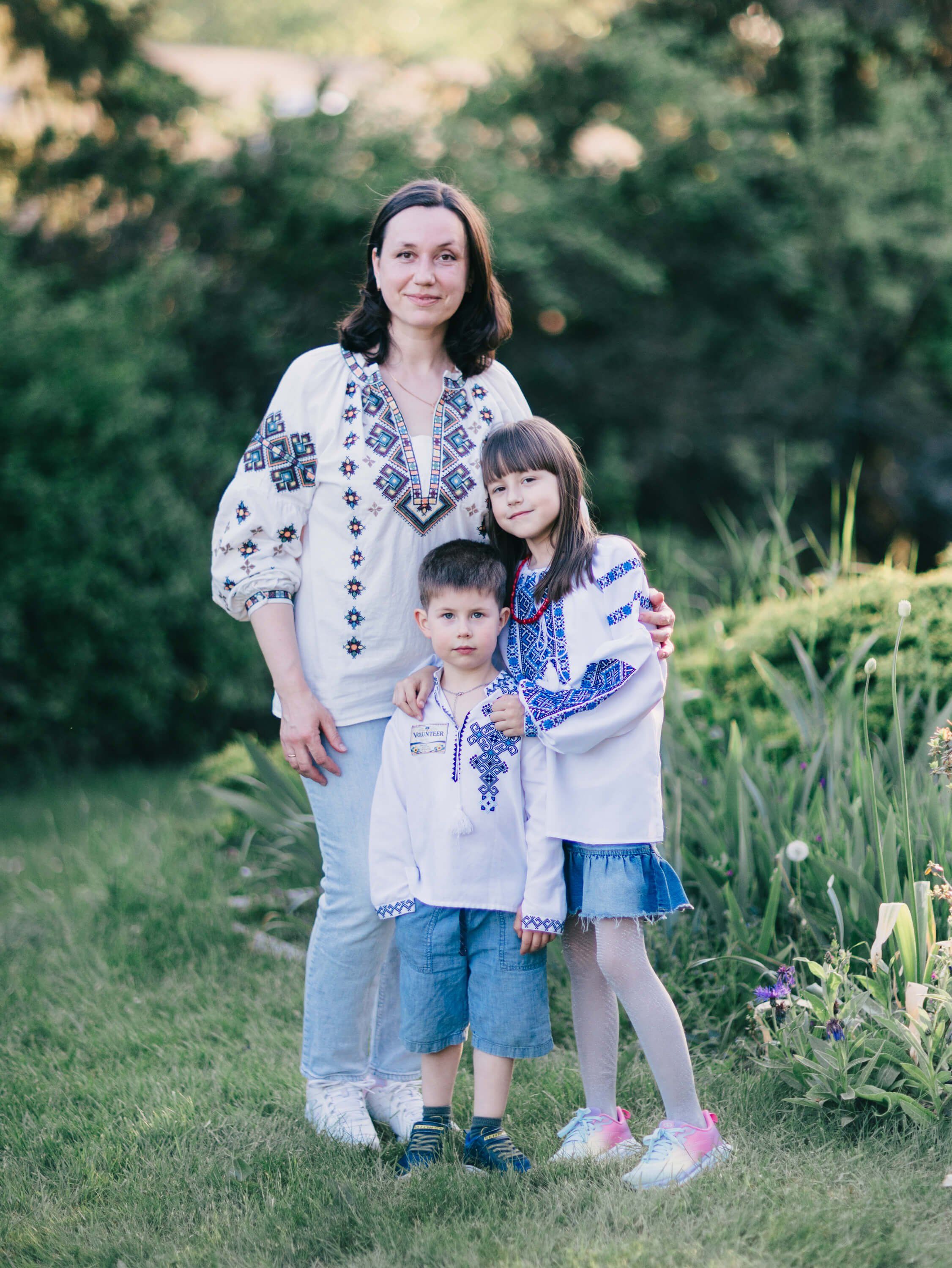 Woman and two children in embroidered Ukrainian clothing standing together in a sunlit garden setting