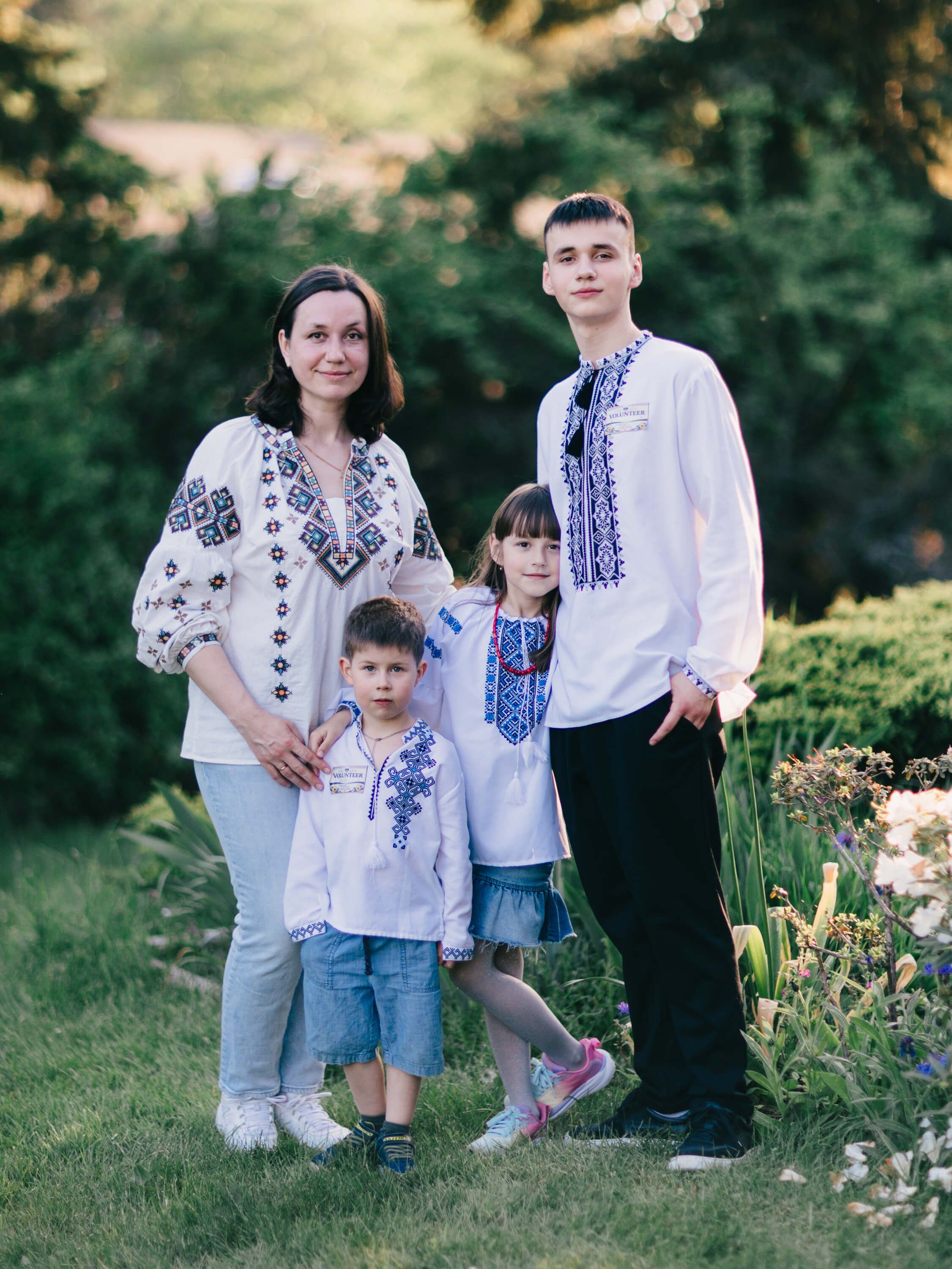 Family of four in coordinating embroidered Ukrainian shirts standing together on a sun-dappled lawn with flowering garden and trees behind them