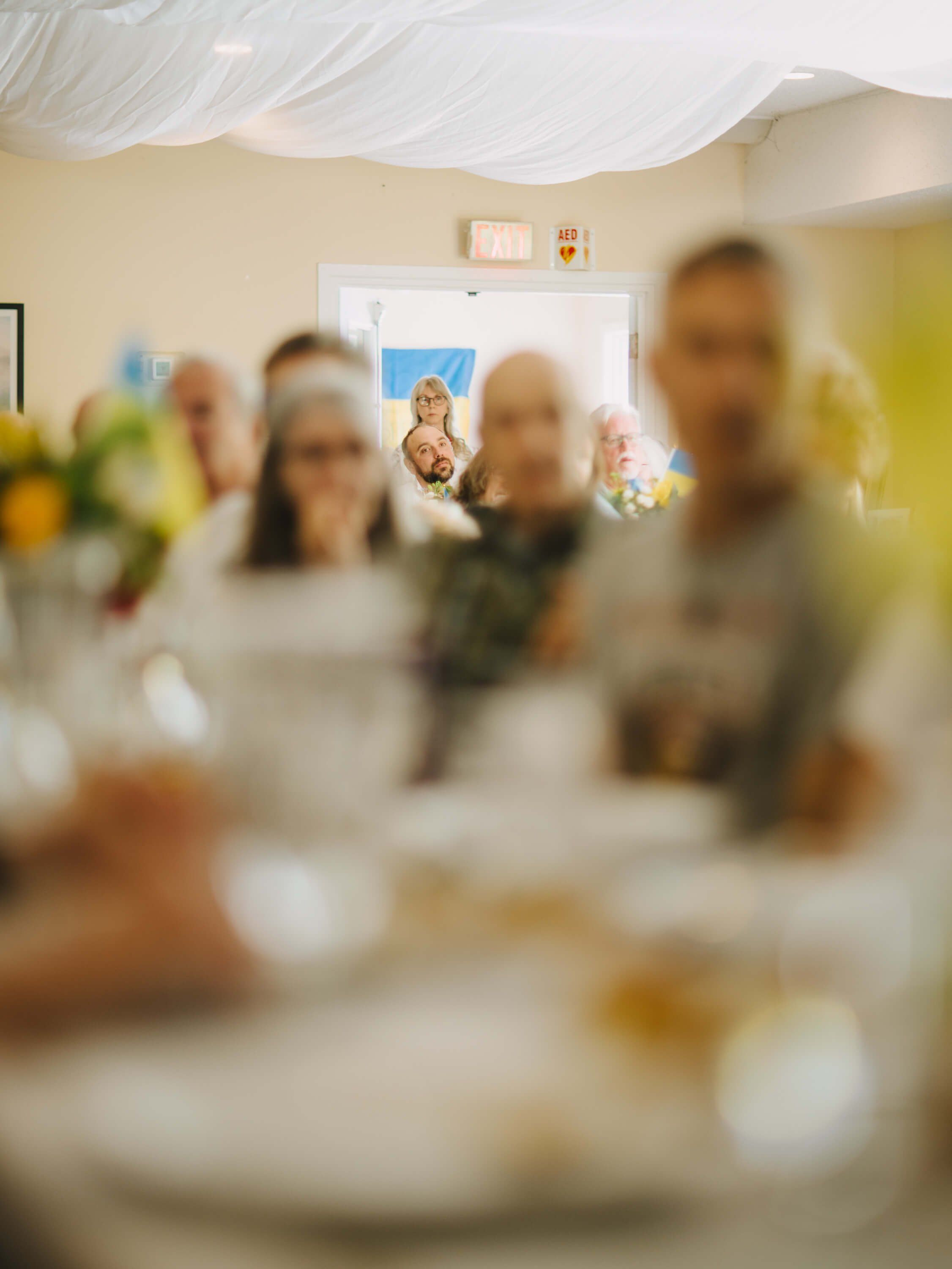 Blurred figures gather in a warmly lit community space with white draped ceiling, a doorway framing the scene beyond