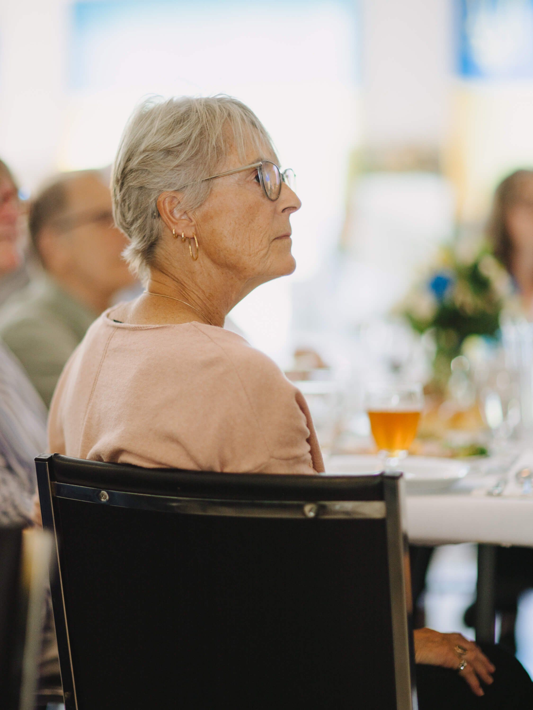 Older woman in glasses and beige top seated at dinner table, listening thoughtfully during the Ukrainian fundraiser event