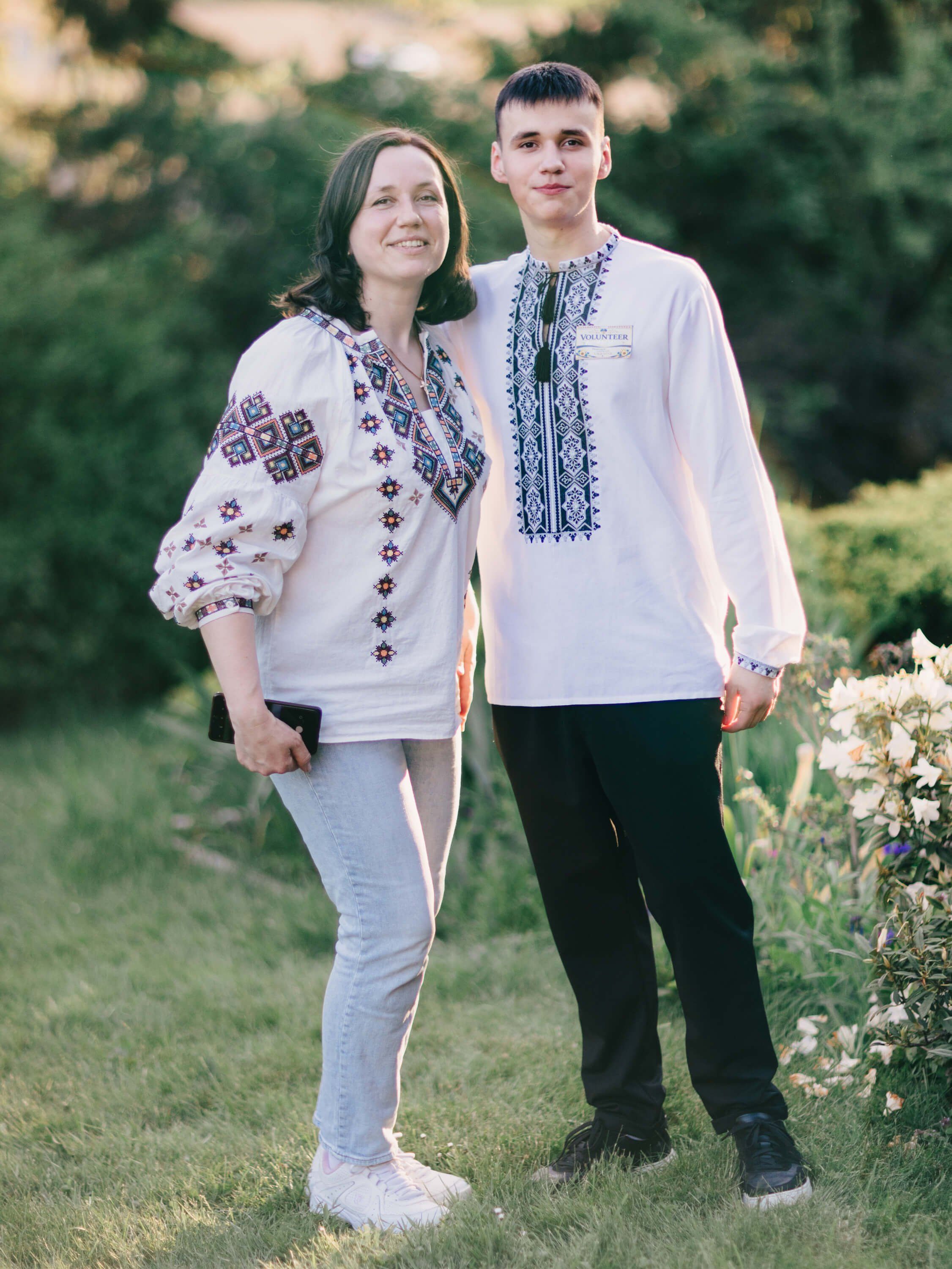 Young couple in matching embroidered white shirts standing on grass at golden hour with green foliage behind them