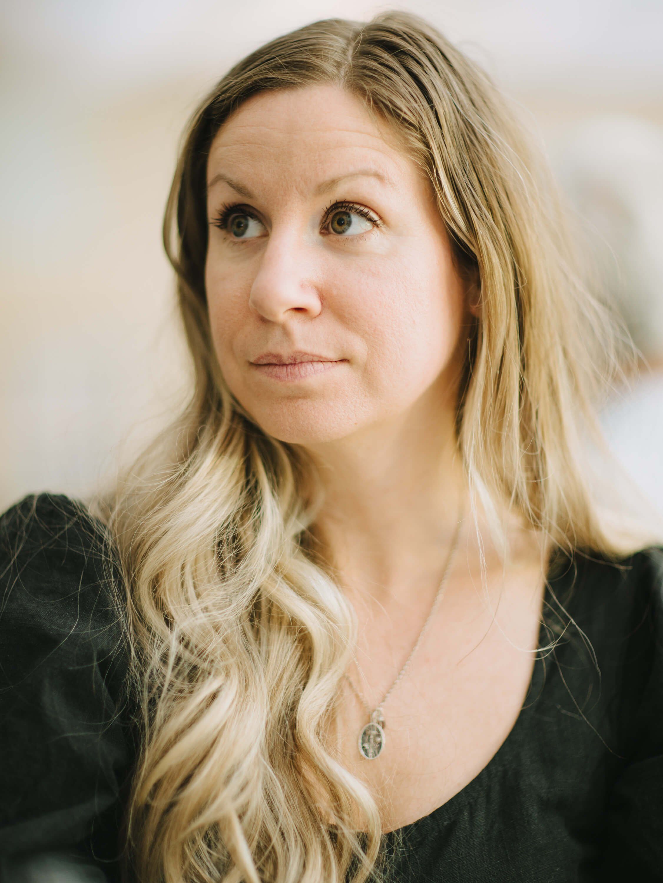 Woman with long blonde hair in black top, soft natural light, gentle expression against blurred background