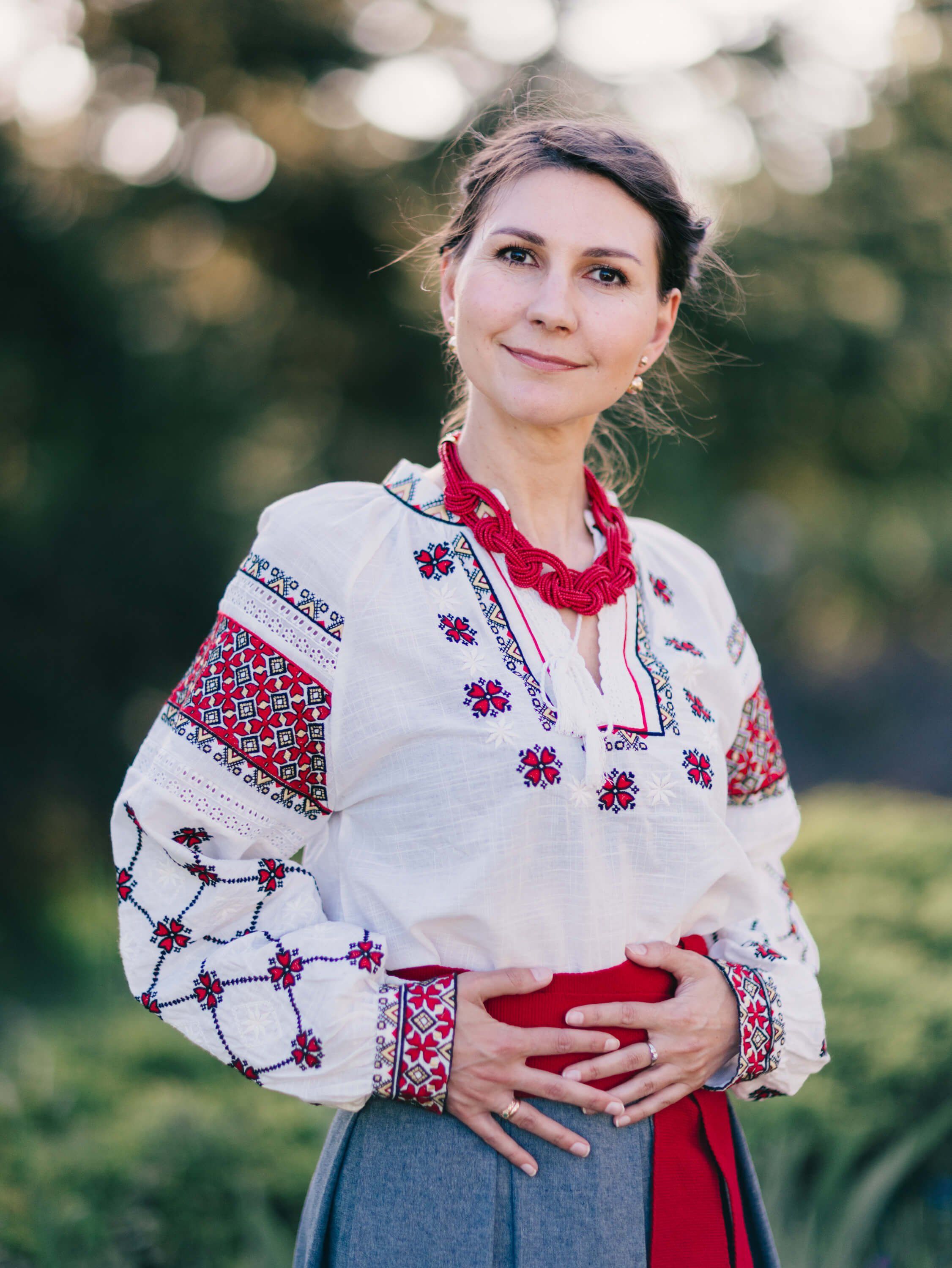 Woman in embroidered Ukrainian blouse and red necklace smiles softly in golden outdoor light, hands clasped at waist