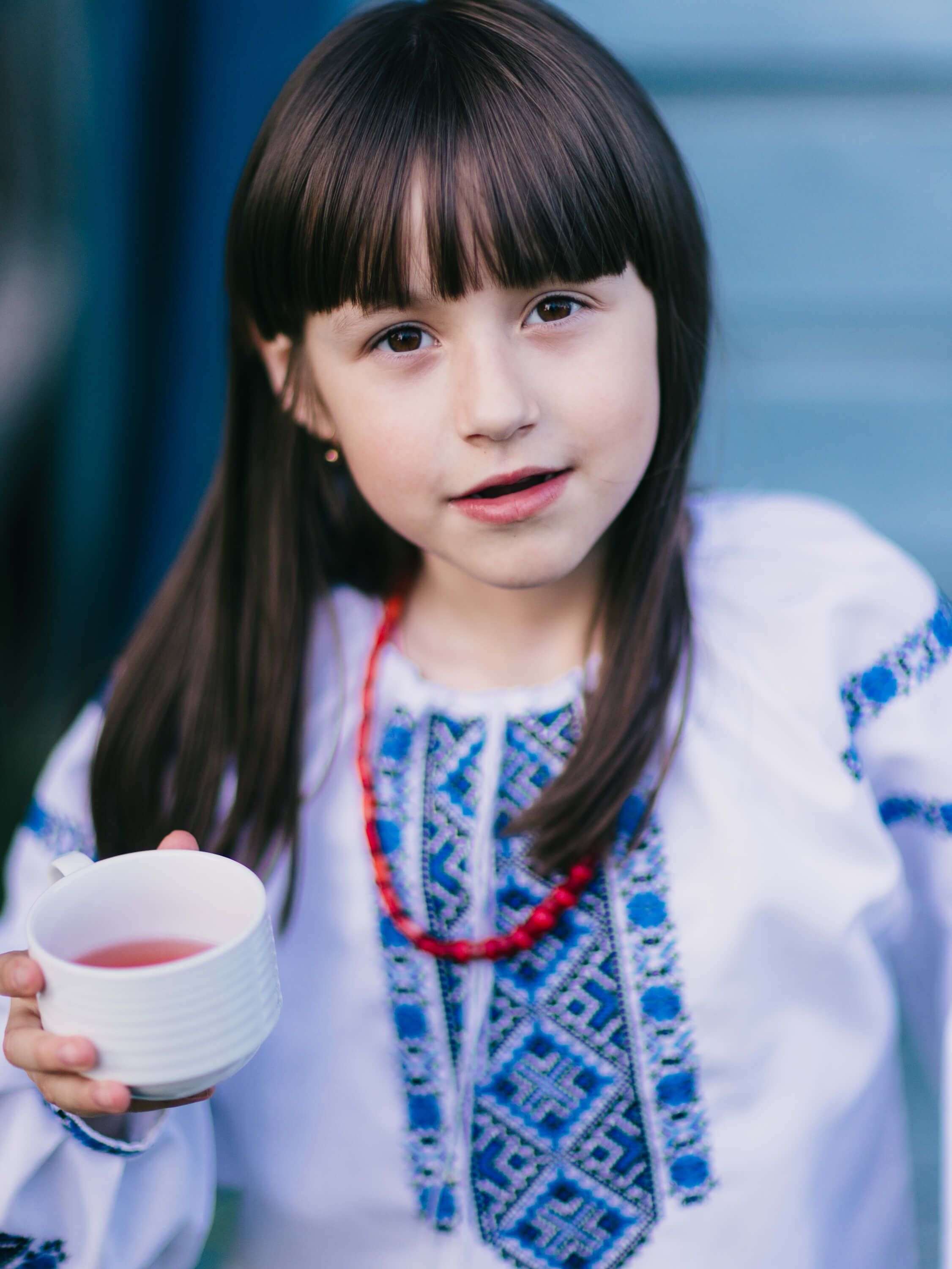 Young girl in embroidered white Ukrainian dress holding a cup of borscht, soft blue background, gentle natural light