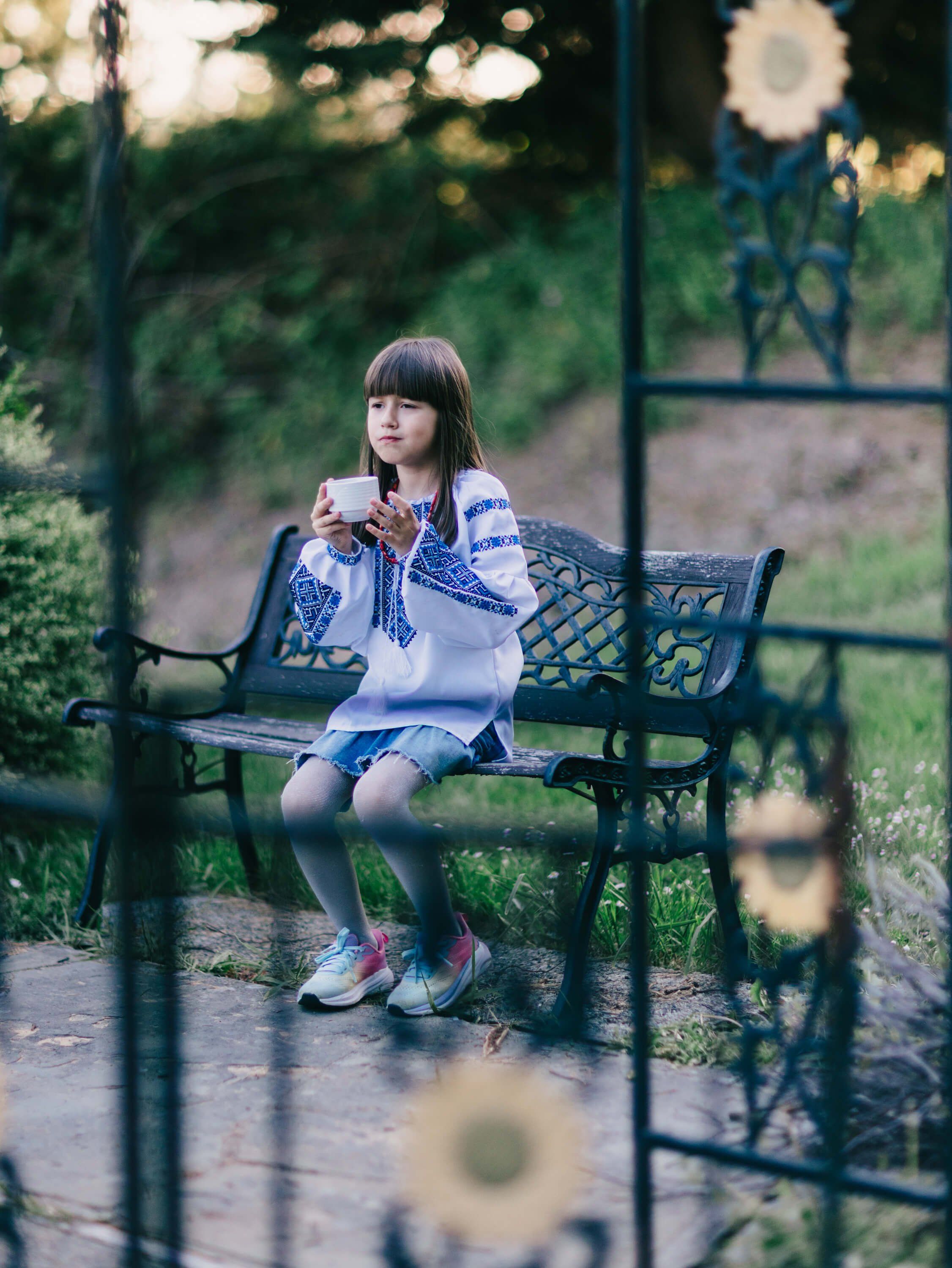 Girl in embroidered Ukrainian shirt seated on bench at golden hour, framed by blurred fence and dandelion bokeh