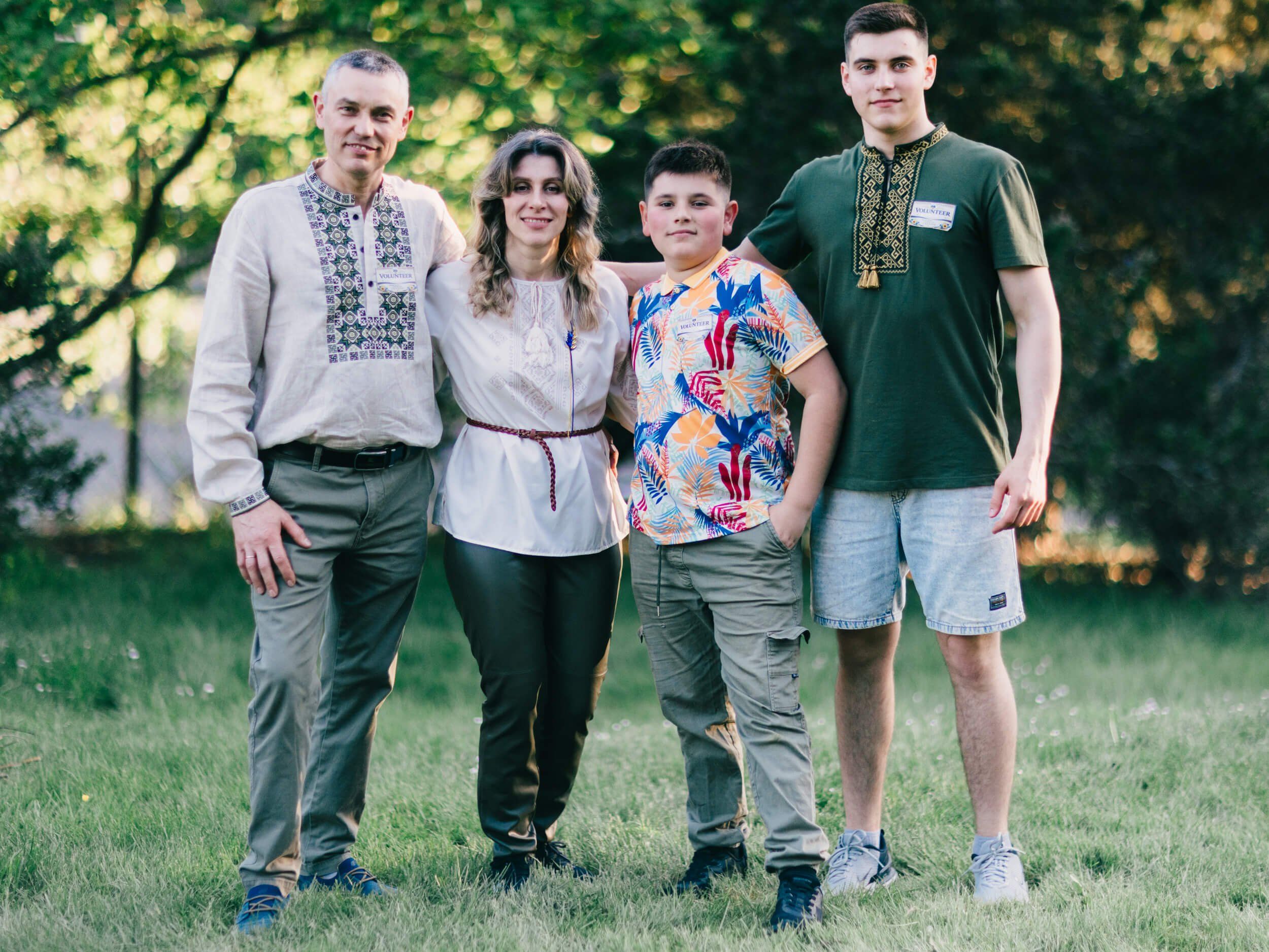 Four people in traditional embroidered shirts stand together on grass with golden-hour trees behind them