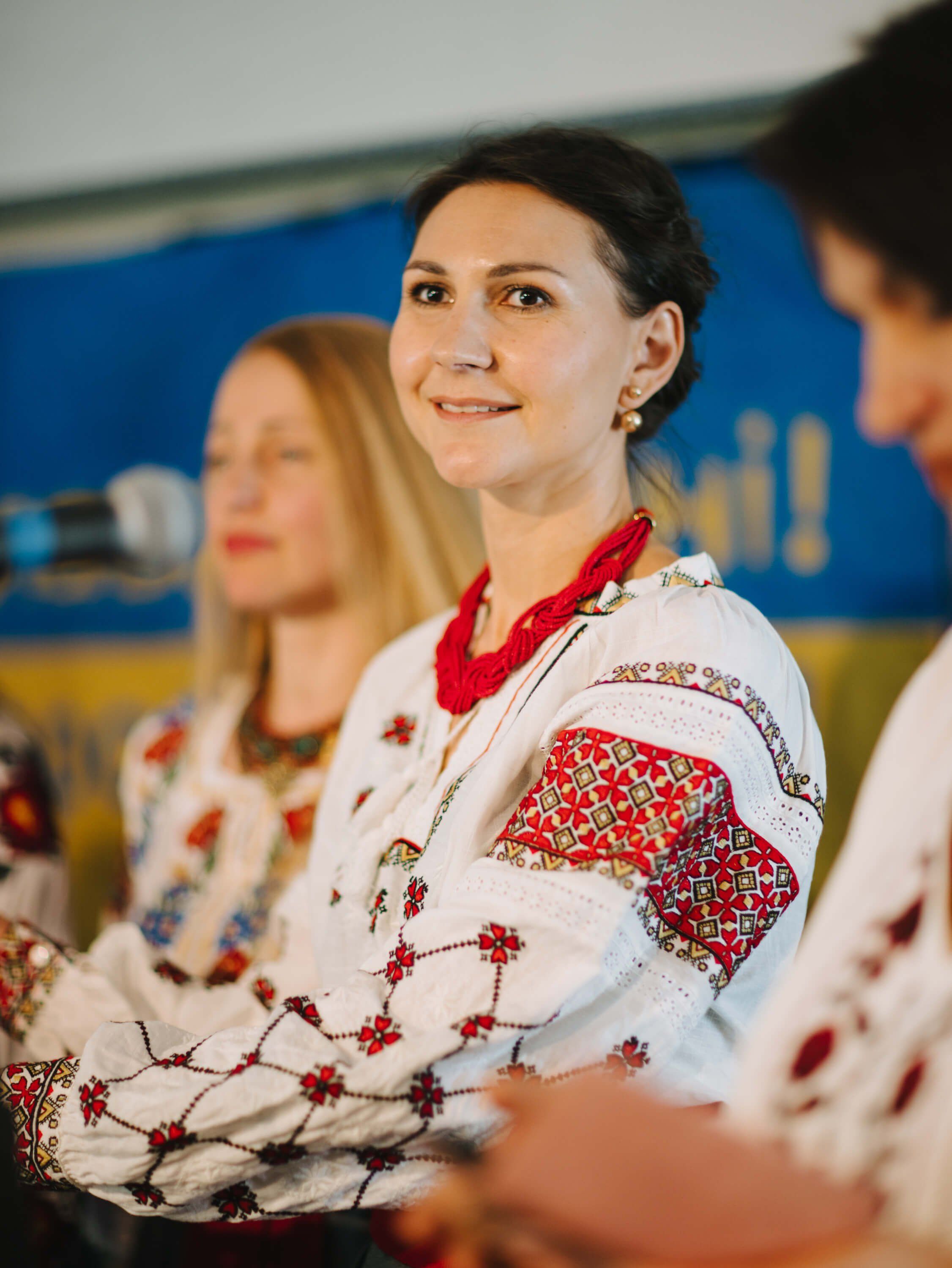 Woman in embroidered red and white Ukrainian dress smiles at Salt Spring Island fundraiser dinner, soft blue backdrop behind