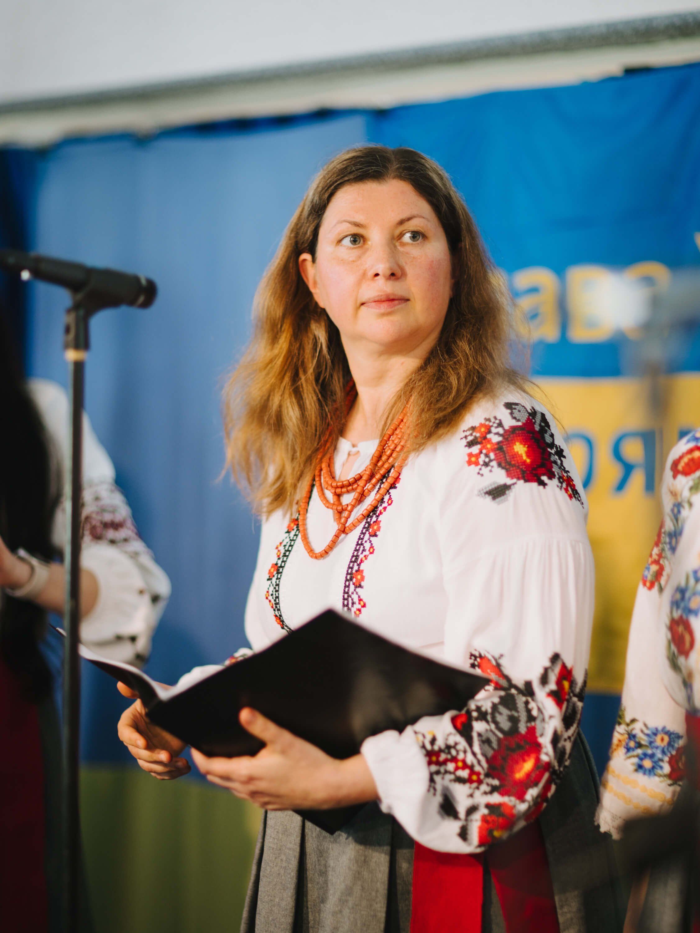 Woman in embroidered Ukrainian dress speaks at microphone during Salt Spring Island fundraiser dinner, holding black folder