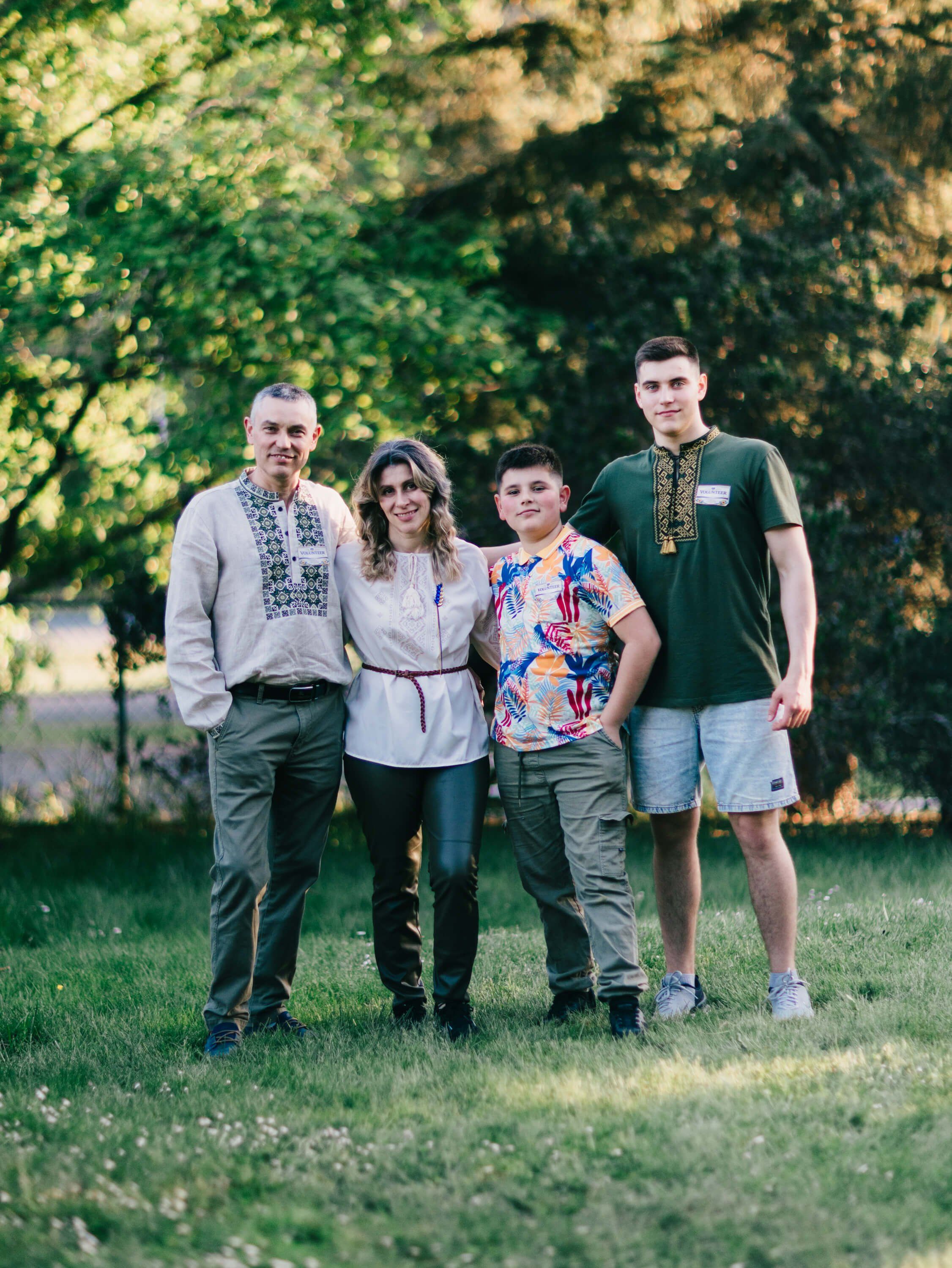 Family of four in embroidered Ukrainian shirts standing on grass beneath dappled tree light at golden hour