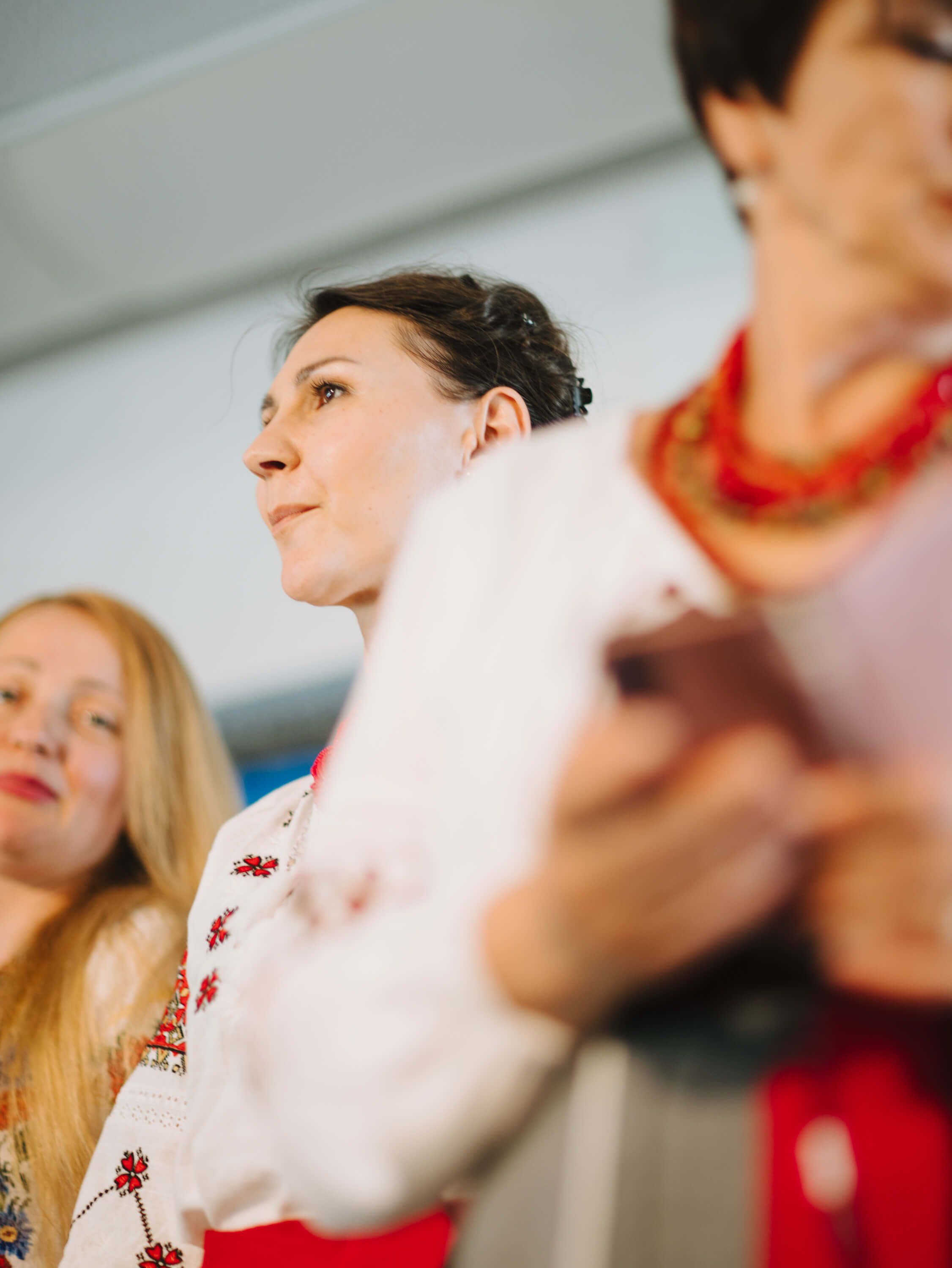 Women in traditional embroidered clothing and beaded necklaces at an indoor fundraiser event, captured in profile with warm natural light