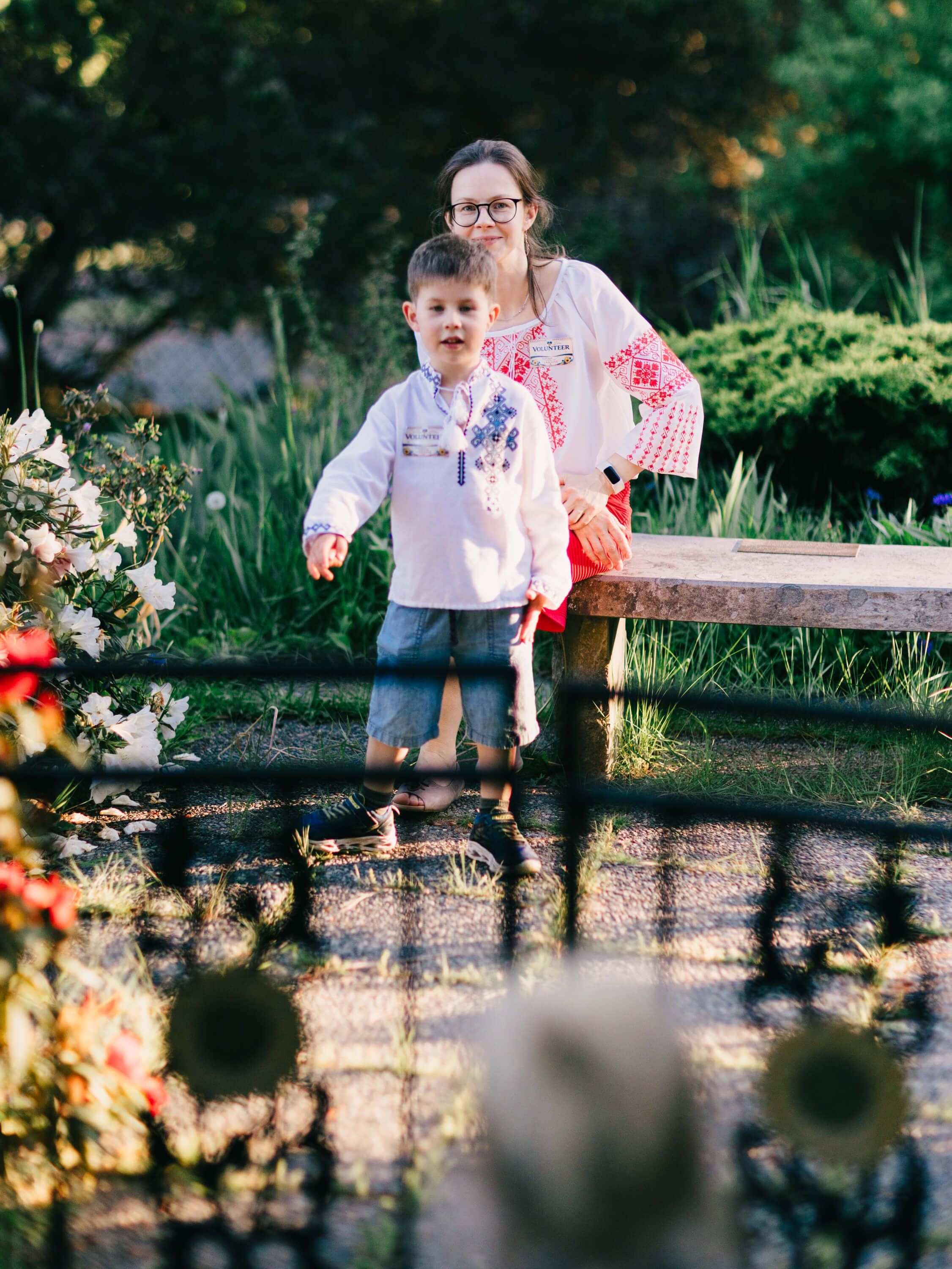 Woman and young child standing by a wooden fence in a sunlit garden with flowering plants and green foliage