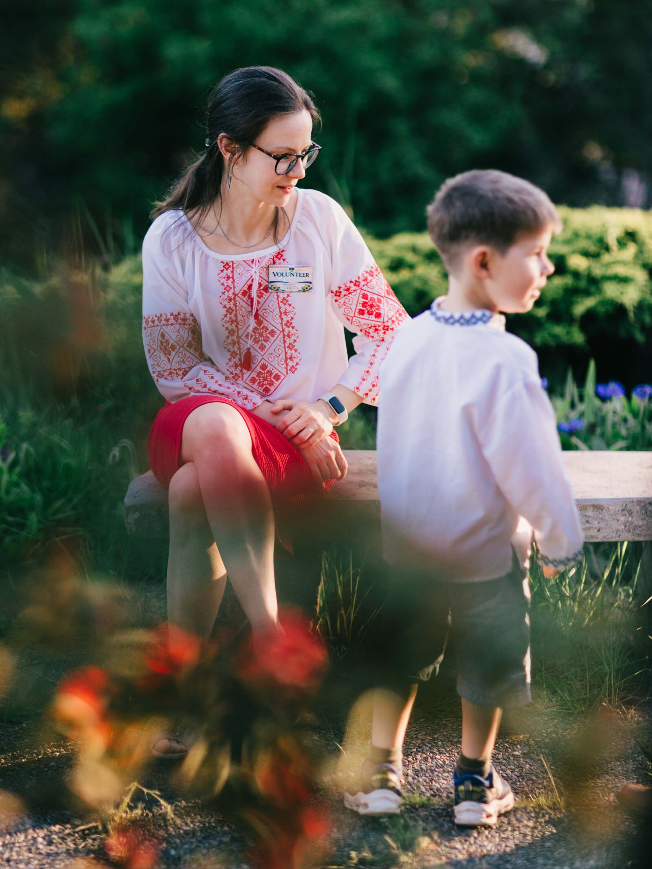 Woman and young boy crouched together by garden flowers, backlit golden hour light filtering through foliage