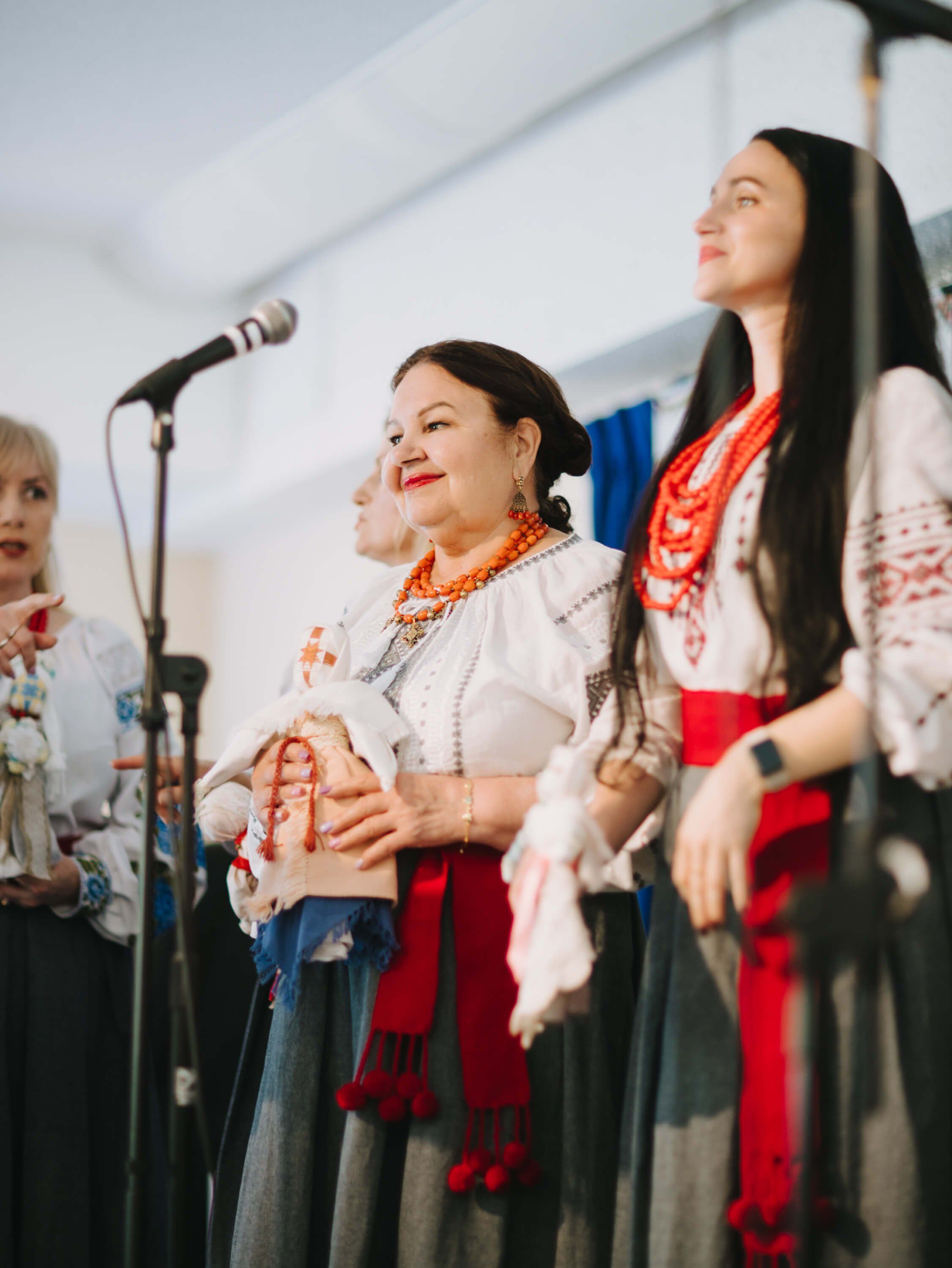 Women in traditional Ukrainian embroidered clothing perform at a microphone during an indoor fundraiser dinner event