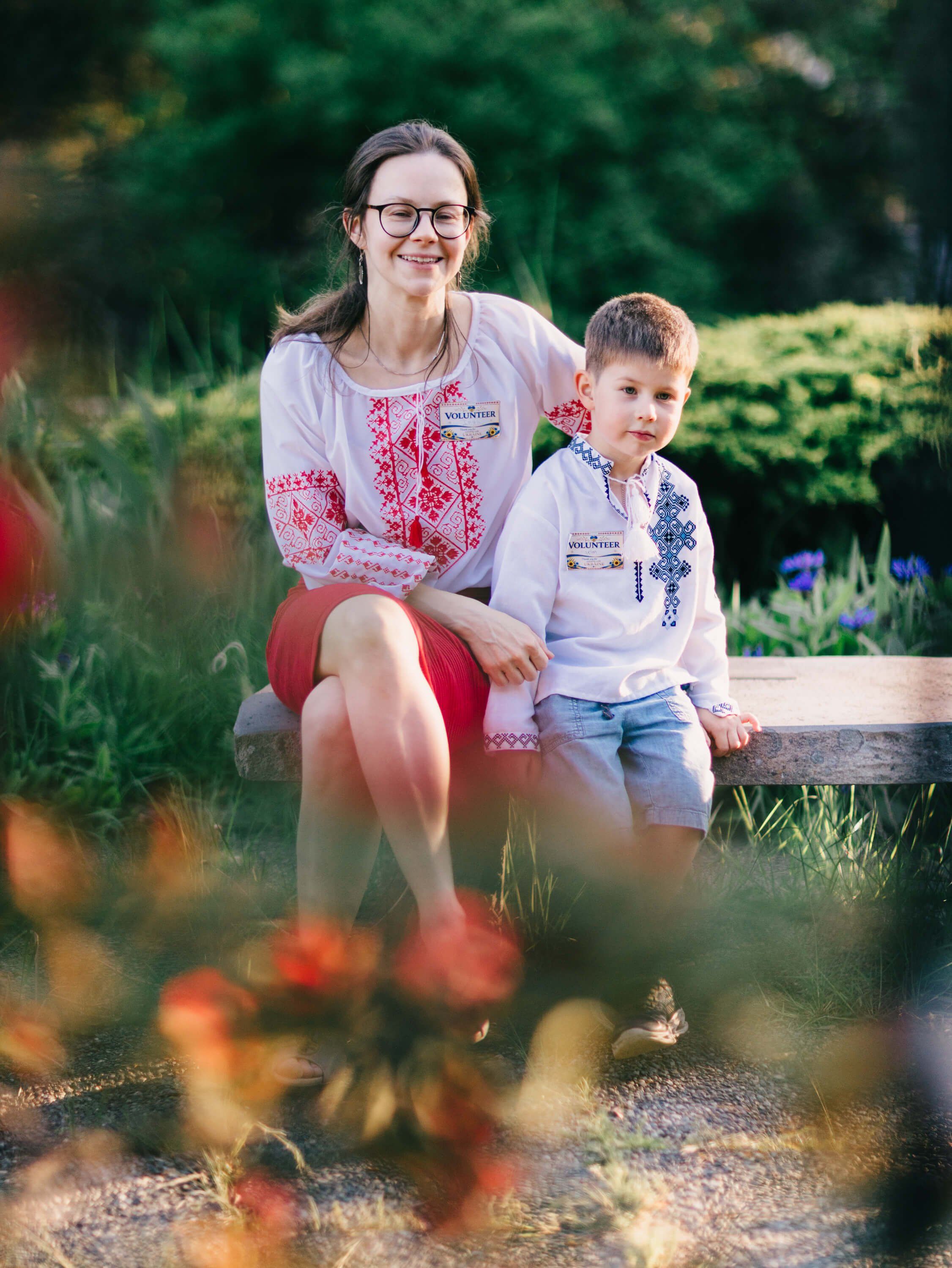 Woman and young boy smiling together in a sunlit garden with flowers and hedges, wearing embroidered traditional clothing