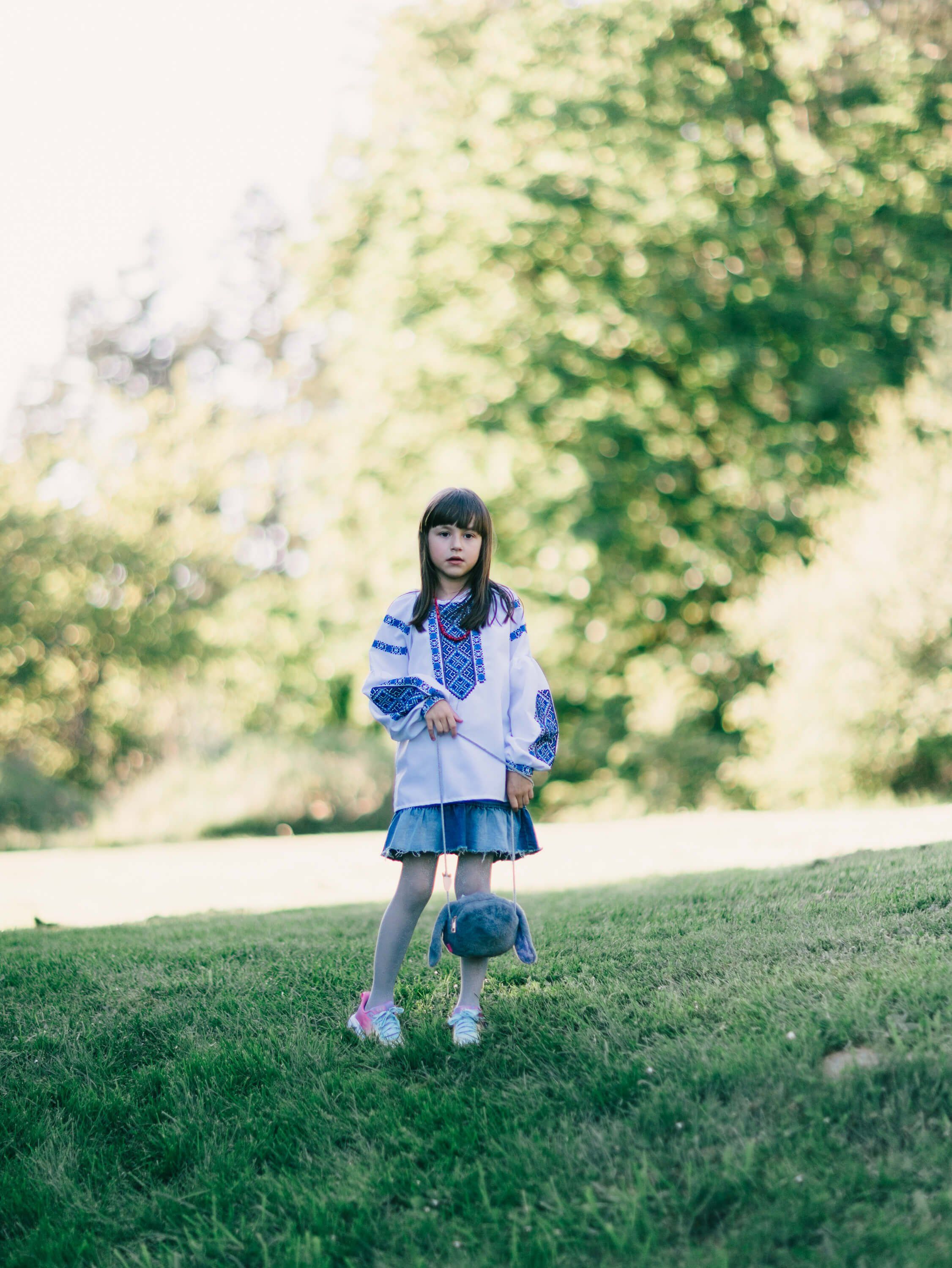 Young girl in blue plaid skirt and white cardigan standing on grass with soft-focus foliage backdrop