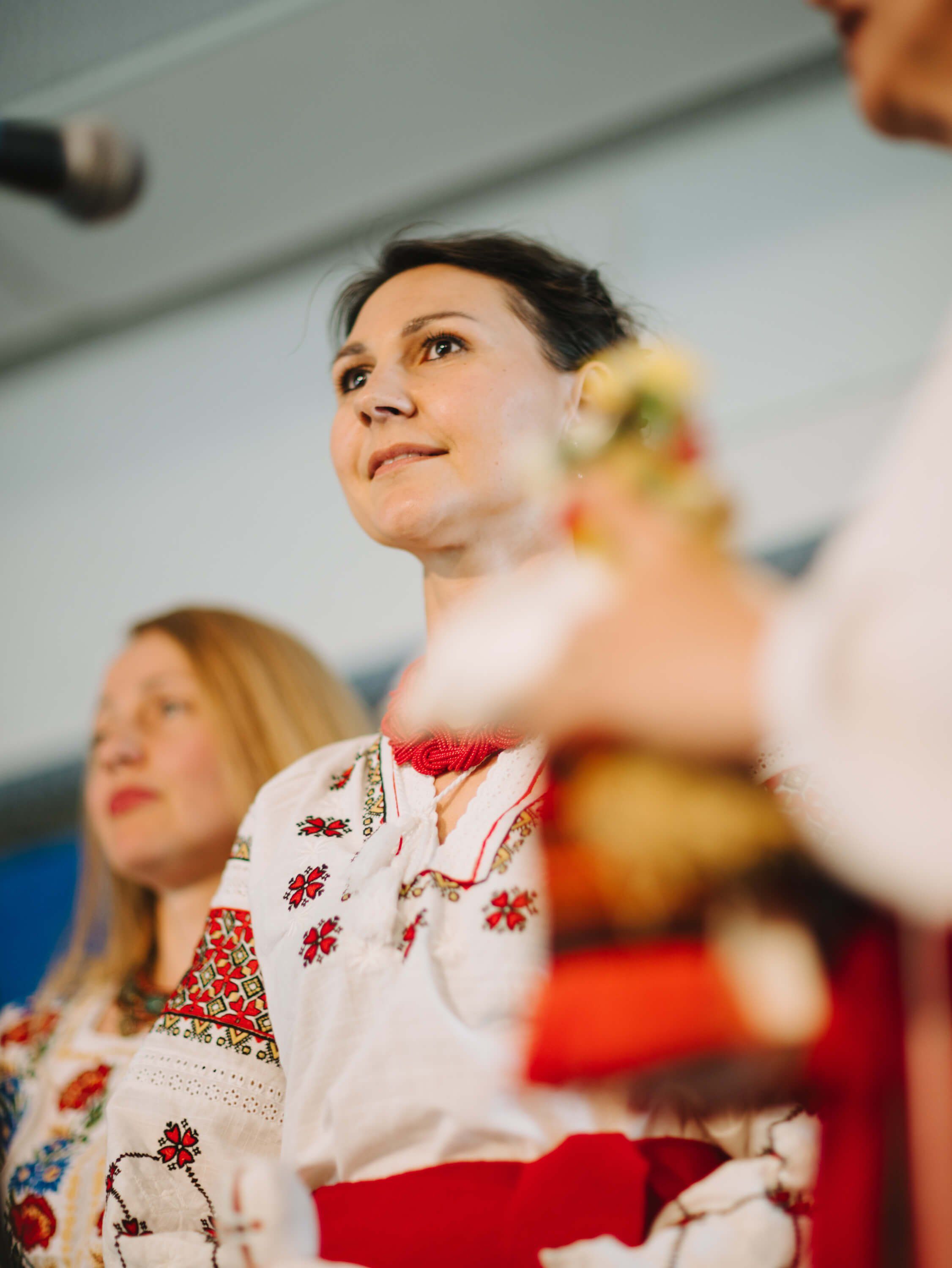 Woman in embroidered white and red traditional Ukrainian dress gazes upward during fundraiser dinner, warm indoor lighting