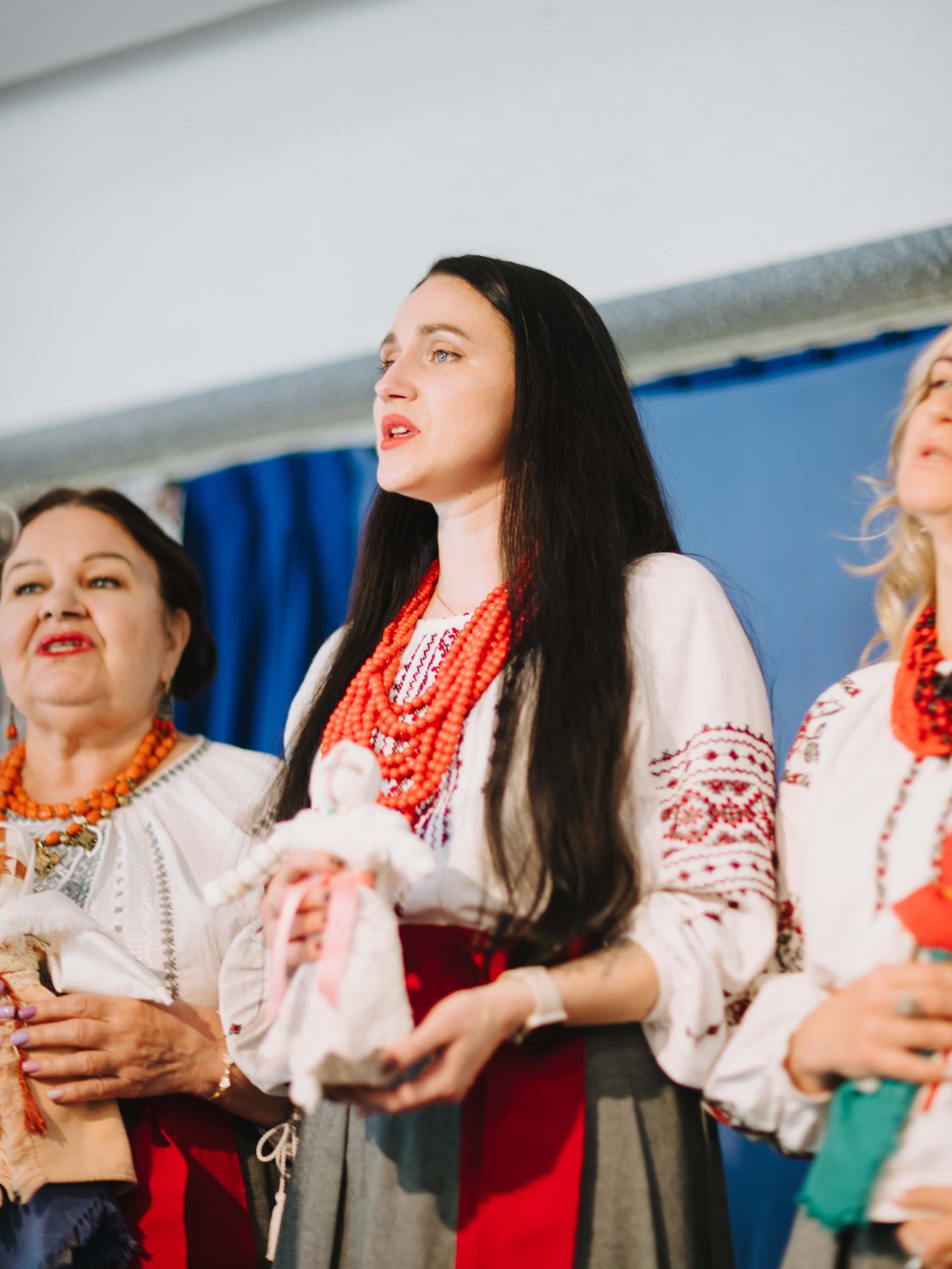 Three women in traditional Ukrainian embroidered white shirts and red beaded necklaces sing together at an indoor fundraiser event