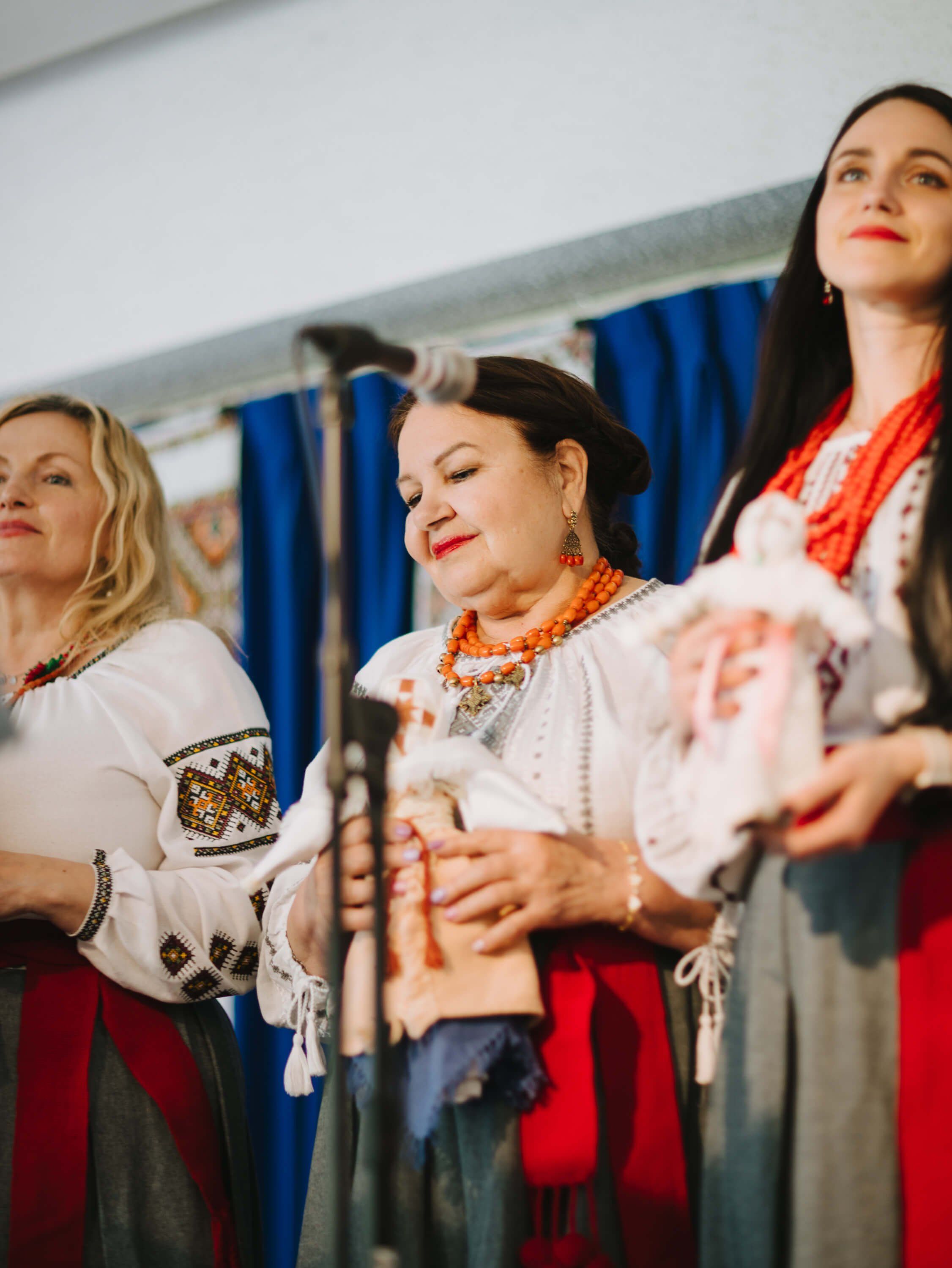 Three women in traditional Ukrainian embroidered shirts and red skirts perform at an indoor fundraiser, smiling warmly under stage lighting with blue curtain…