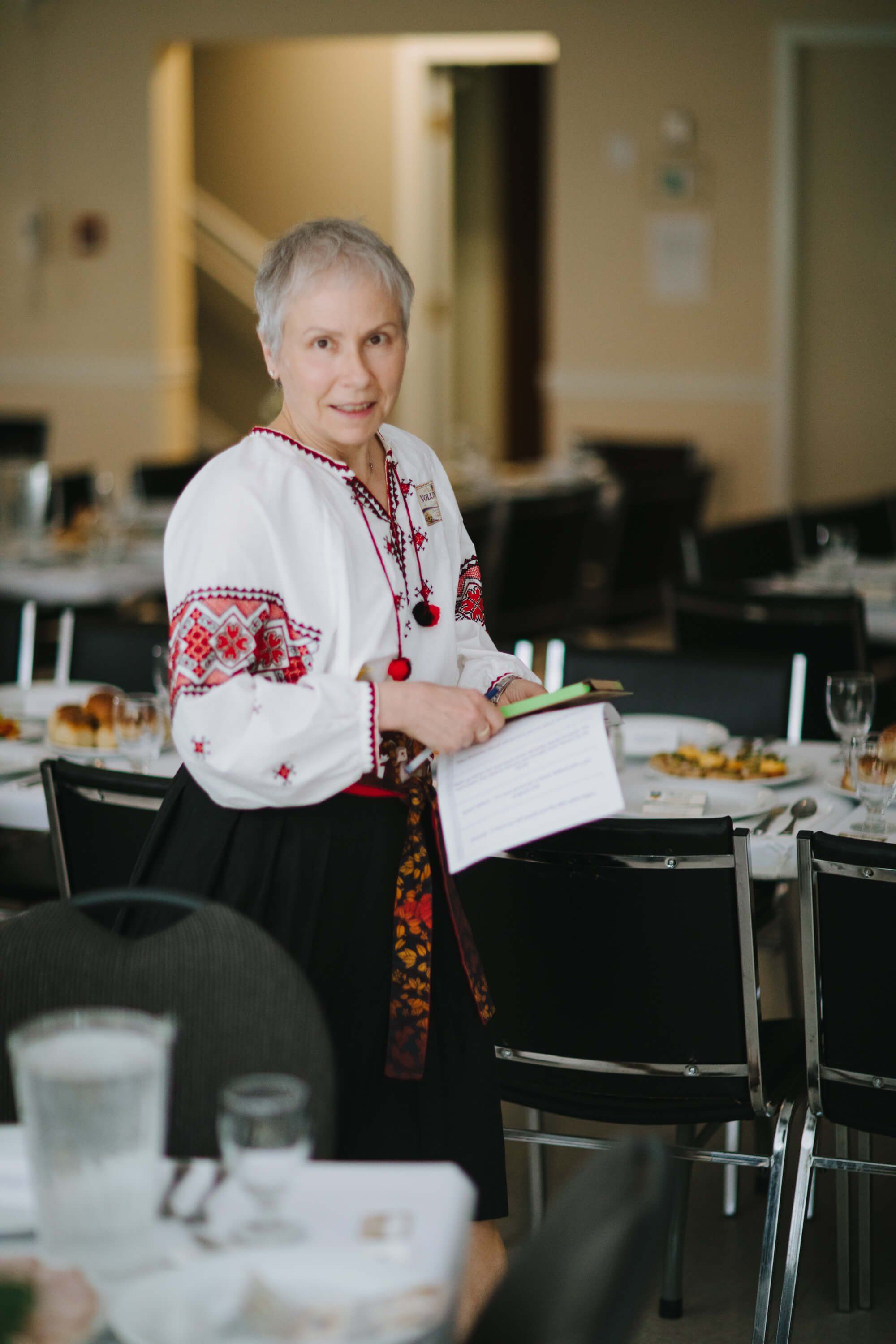 Woman in embroidered white vyshyvanka and red skirt holds a clipboard while standing in a warmly lit dining room during the fundraiser event