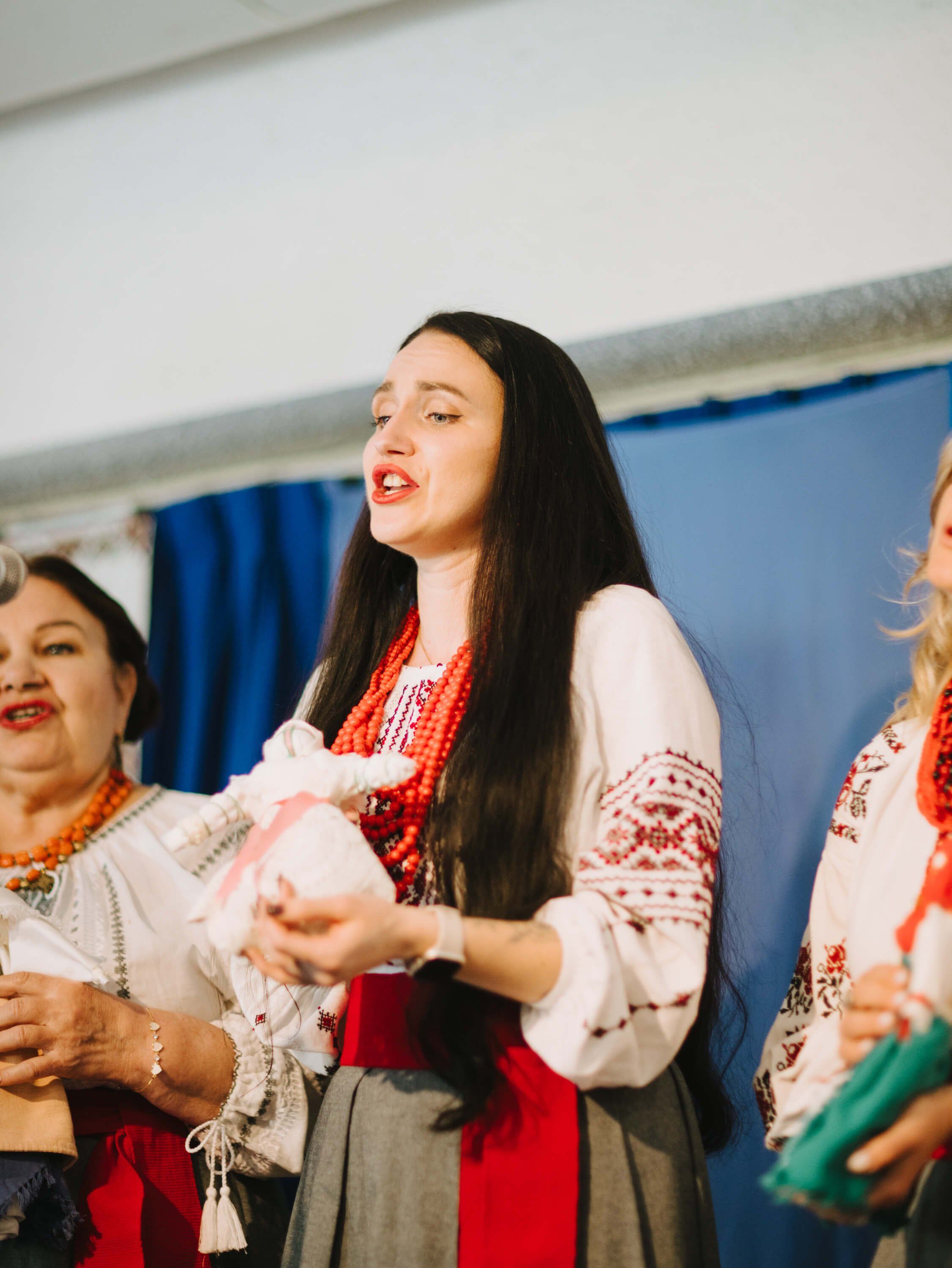 Woman in traditional Ukrainian embroidered dress speaks expressively at indoor fundraiser event beside other performers