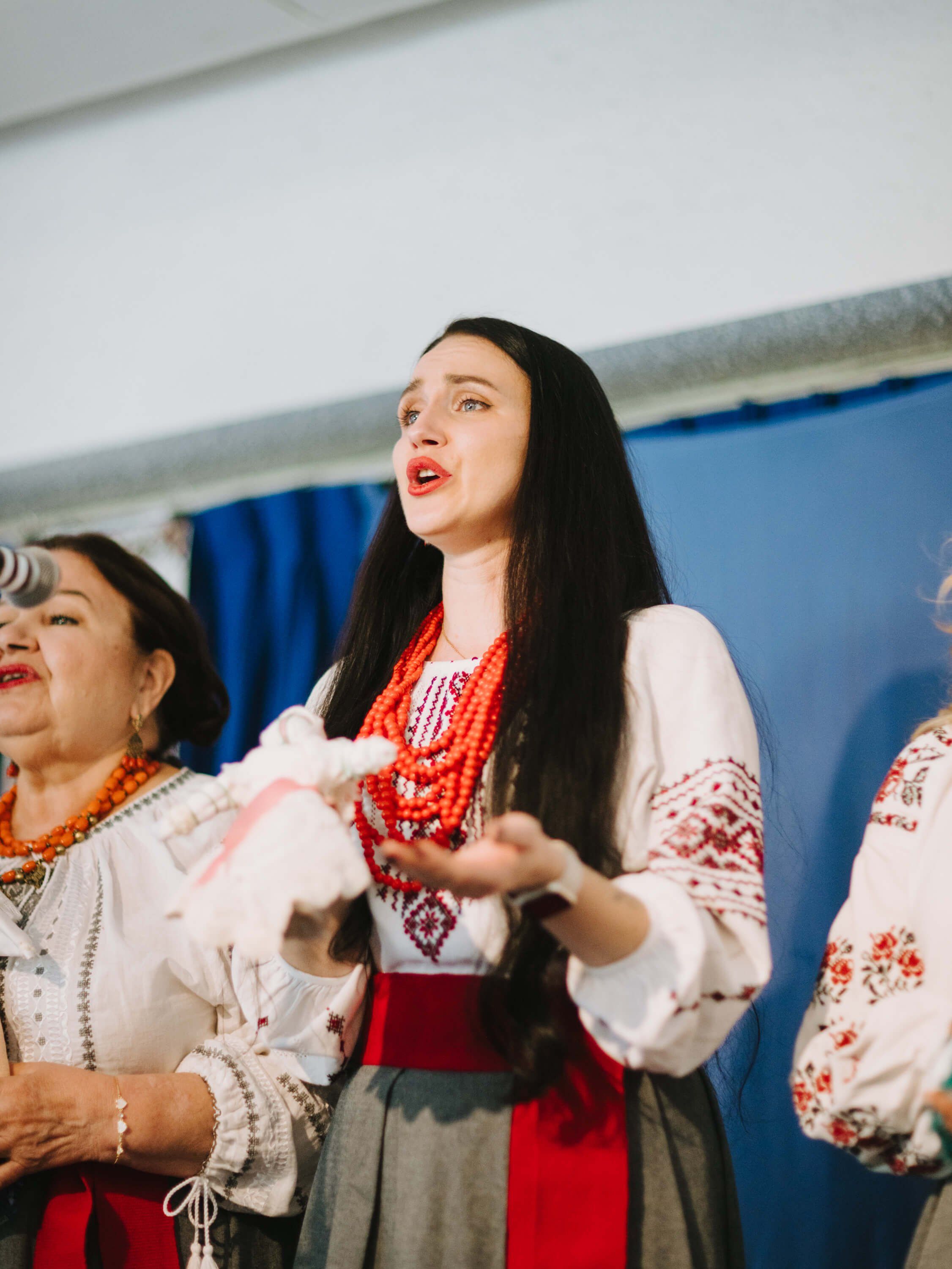 Woman in traditional Ukrainian embroidered dress and red beaded necklace singing with others indoors