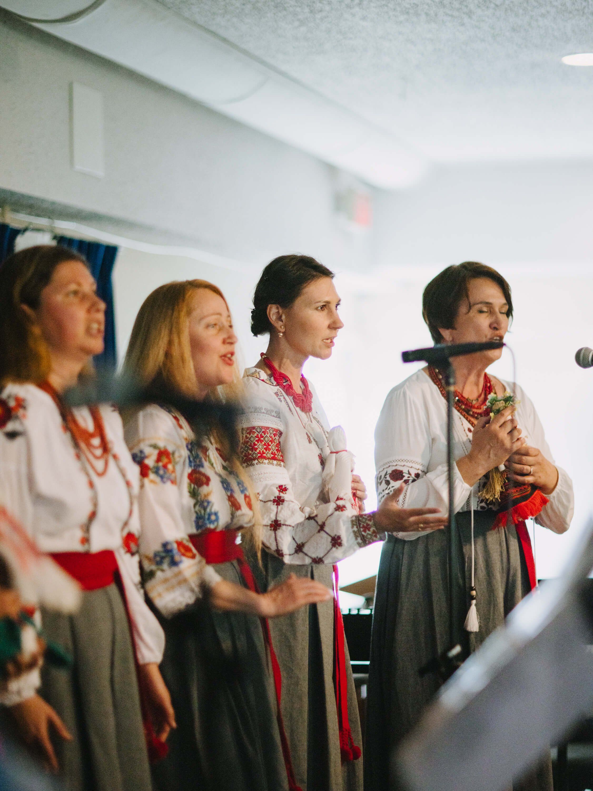 Four women in embroidered Ukrainian folk dresses perform at a microphone indoors, bathed in warm institutional lighting