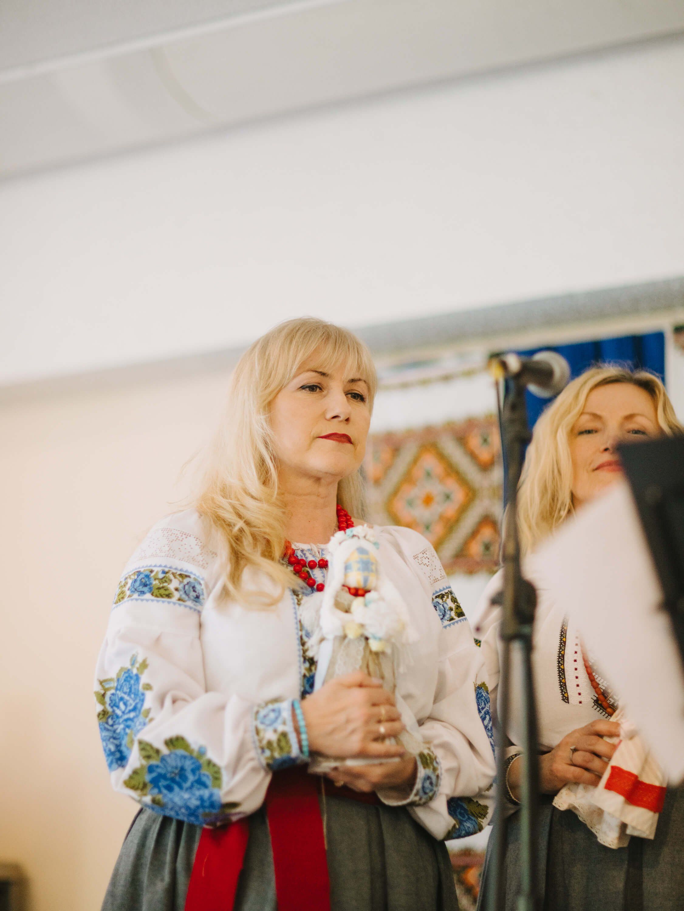 Woman in embroidered Ukrainian blouse and red skirt holds candle at fundraiser dinner, her reflection visible in mirror beside her
