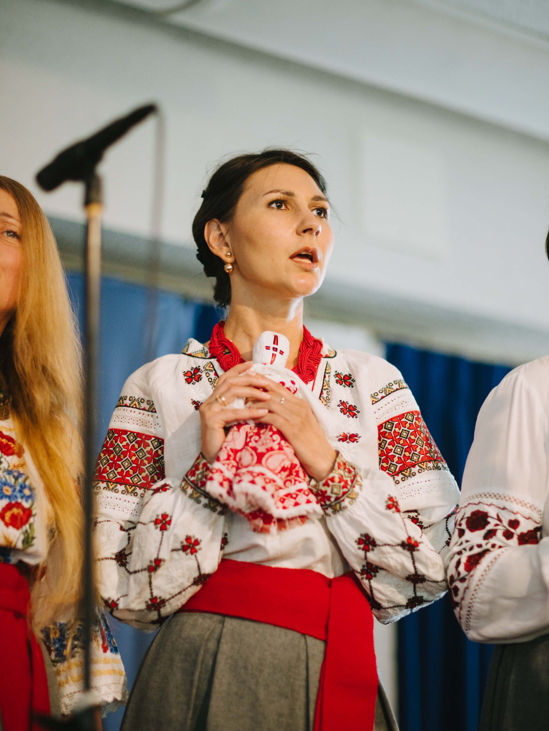 Woman in embroidered Ukrainian vyshyvanka gazes upward while holding a decorative heart, mid-performance at indoor fundraiser dinner