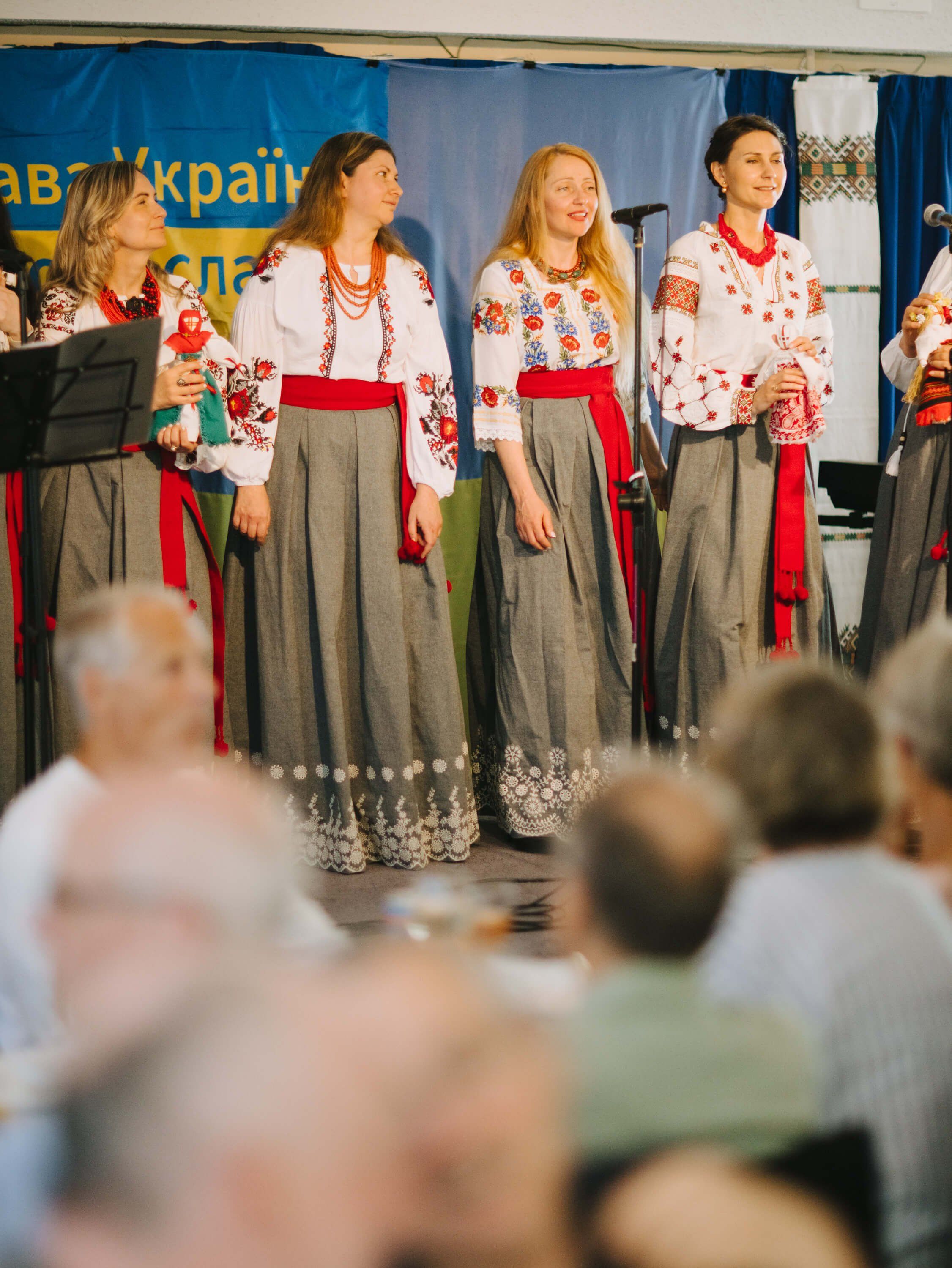 Young women in traditional Ukrainian embroidered blouses and long skirts perform on stage before an audience at an indoor fundraiser dinner