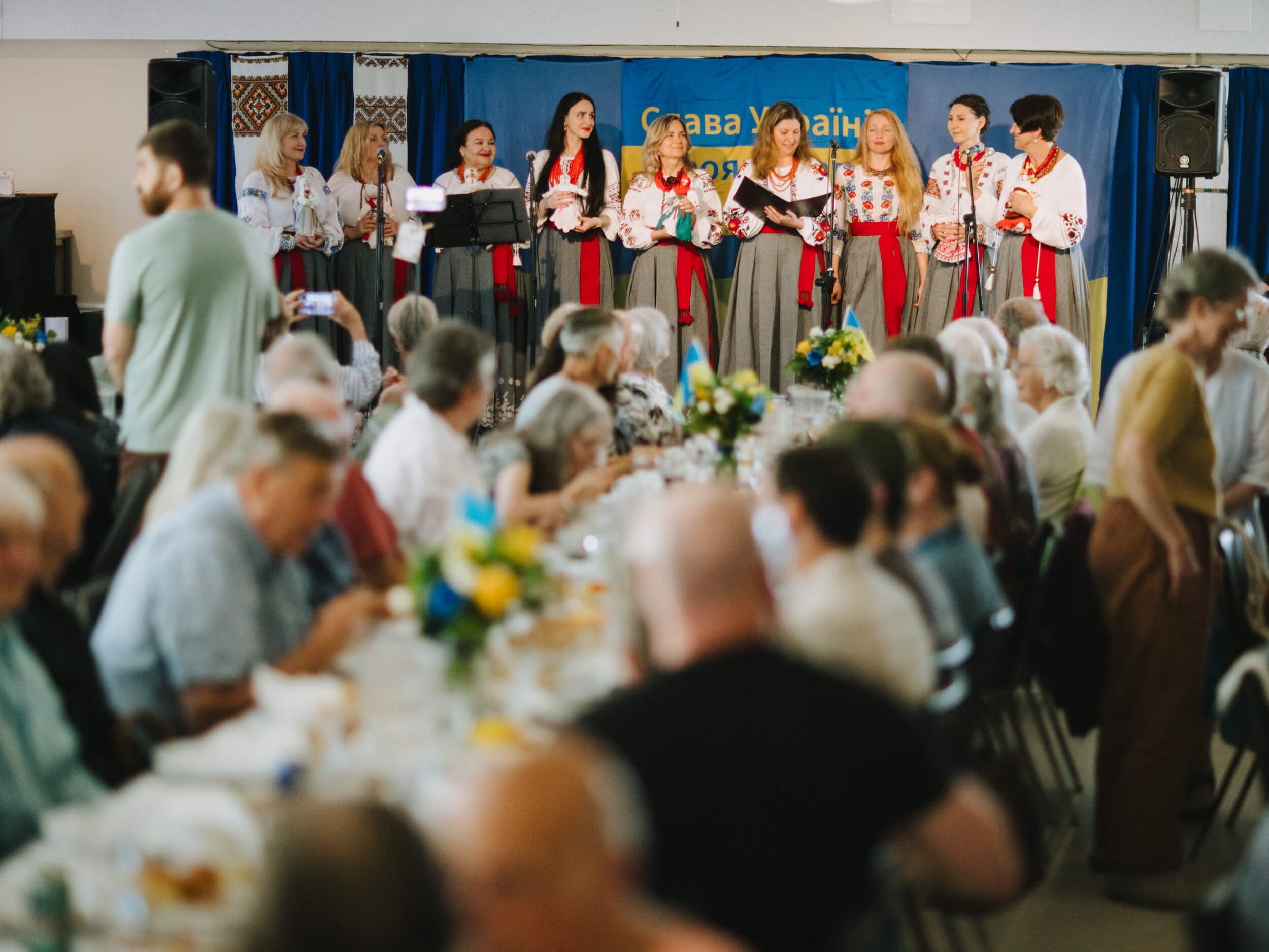 Performers in traditional Ukrainian embroidered clothing stand on stage before a seated audience enjoying dinner at a community fundraiser event
