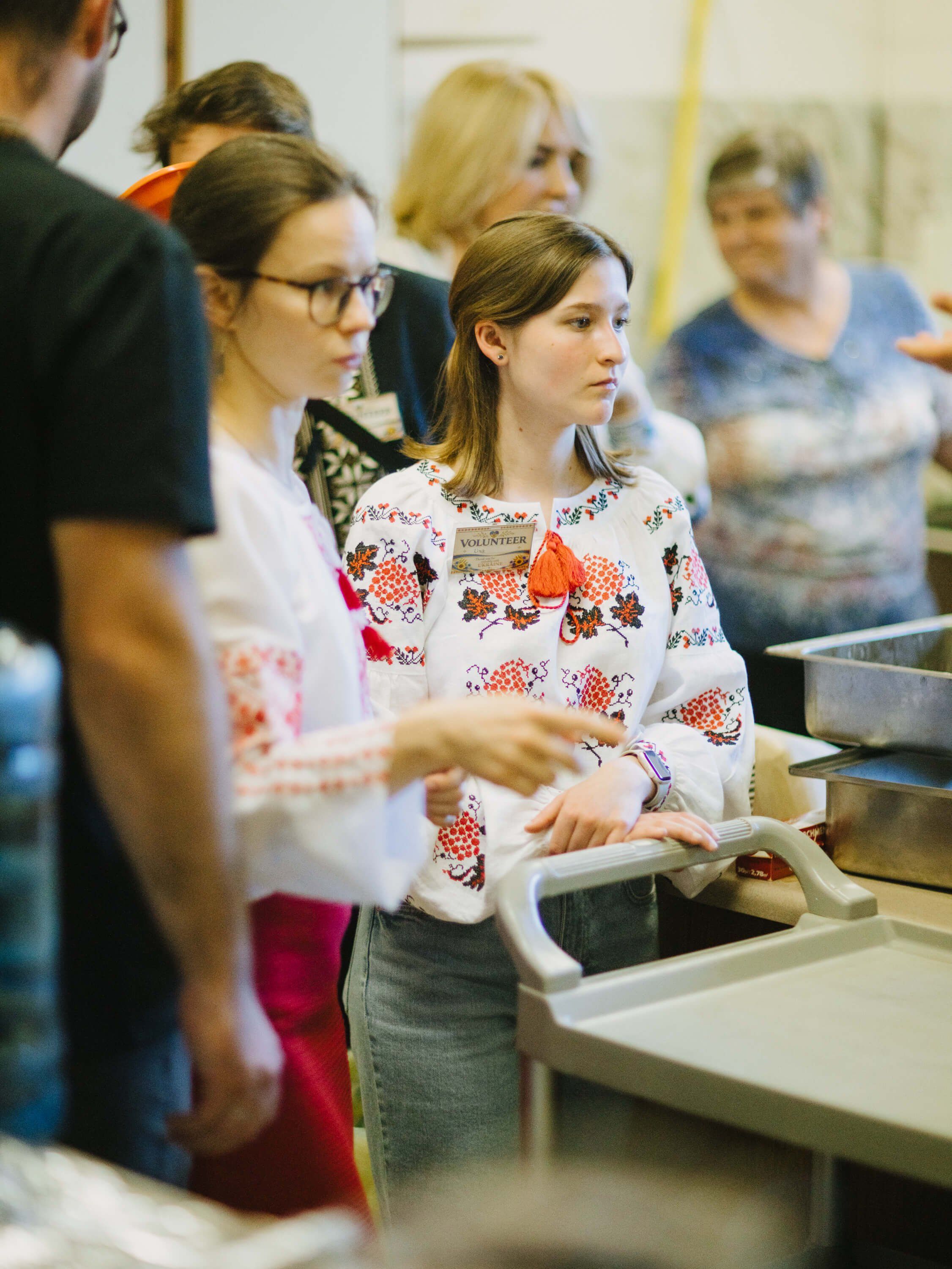 Young woman in floral sweater serves food at community dinner event, surrounded by attendees in warm indoor setting