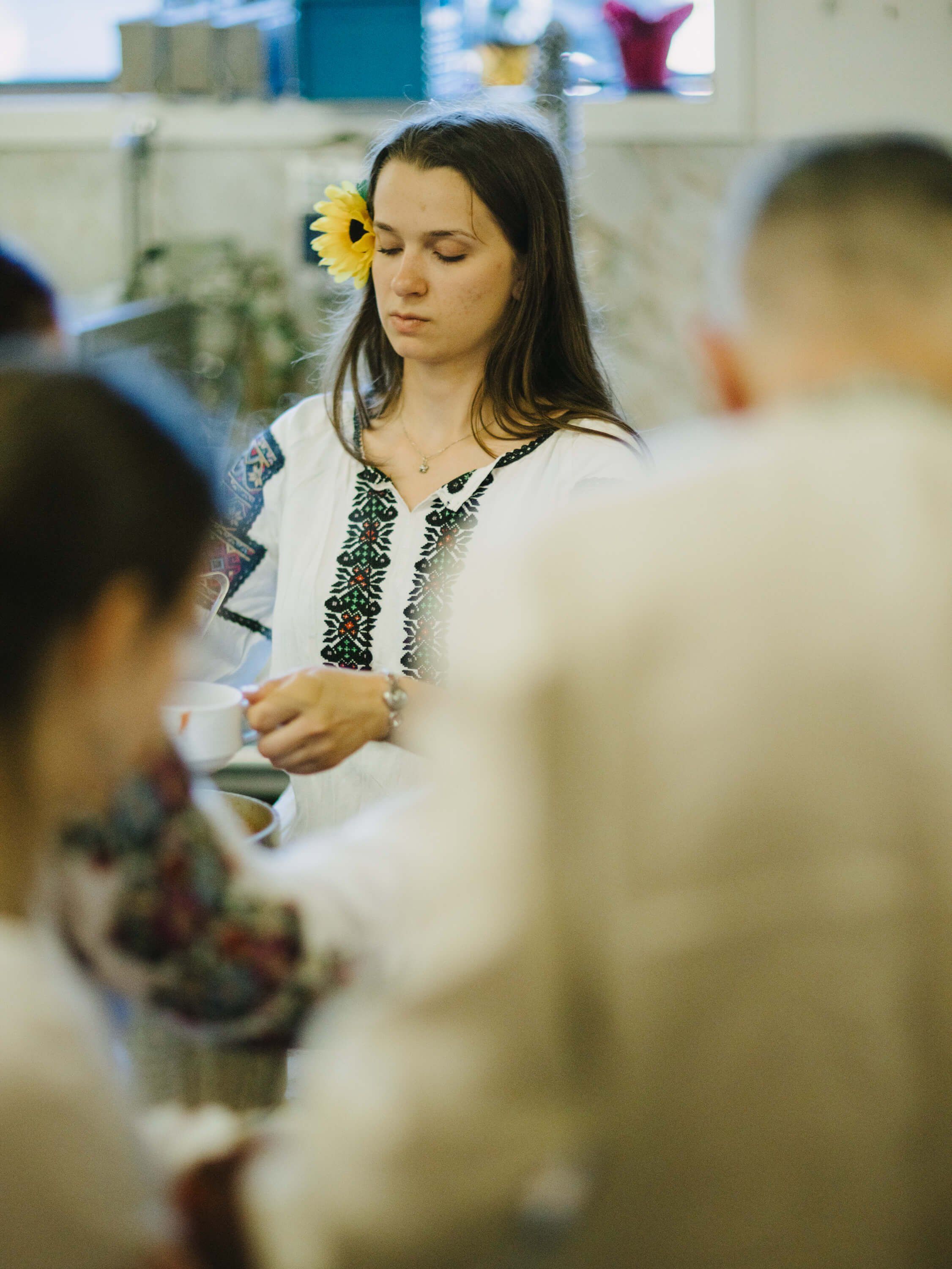 Woman in embroidered white blouse with yellow flower in dark hair serves guests at Ukrainian fundraiser dinner