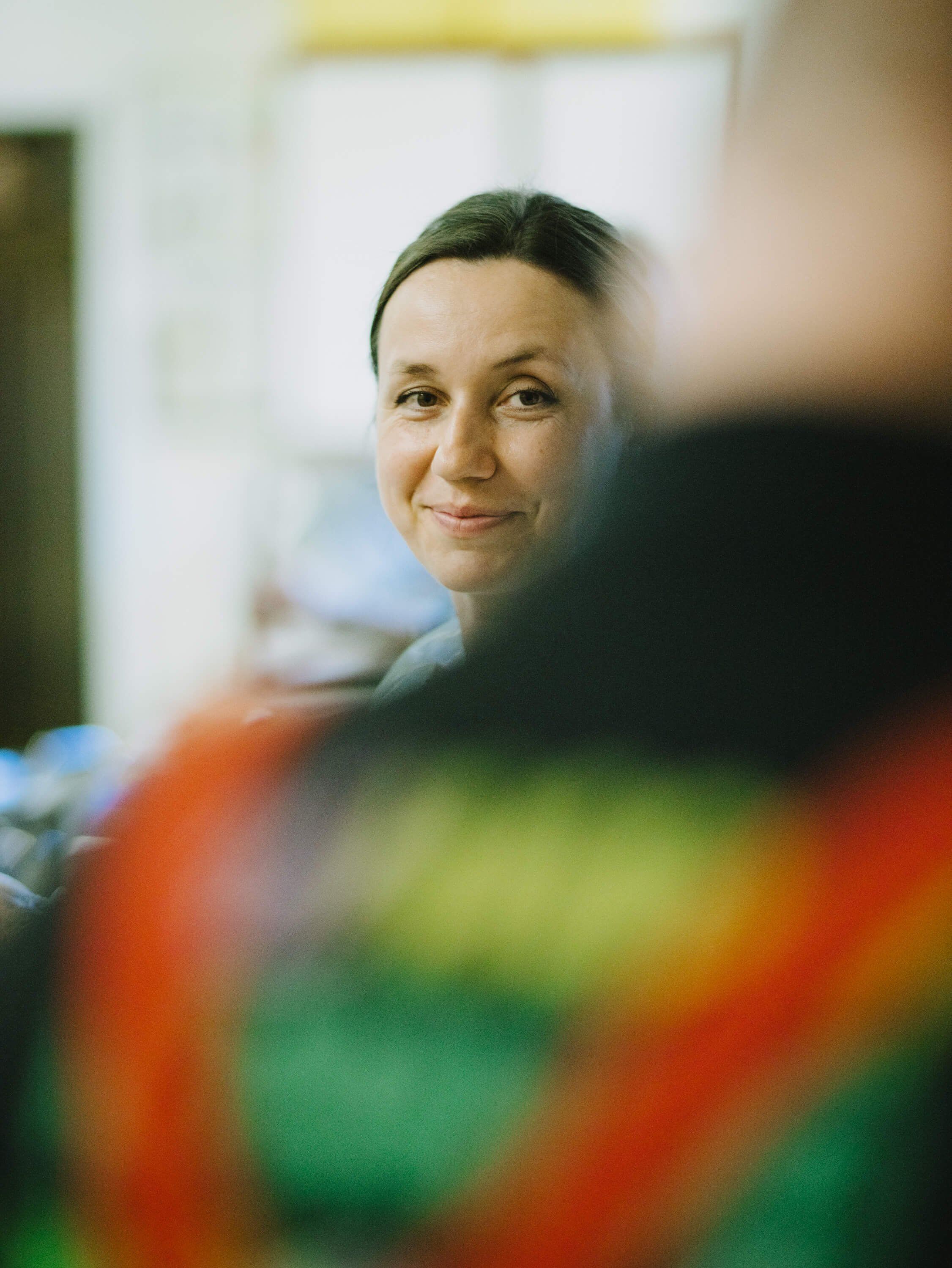 Woman smiling warmly at camera indoors, colorful blurred object in foreground, soft natural light from window behind her