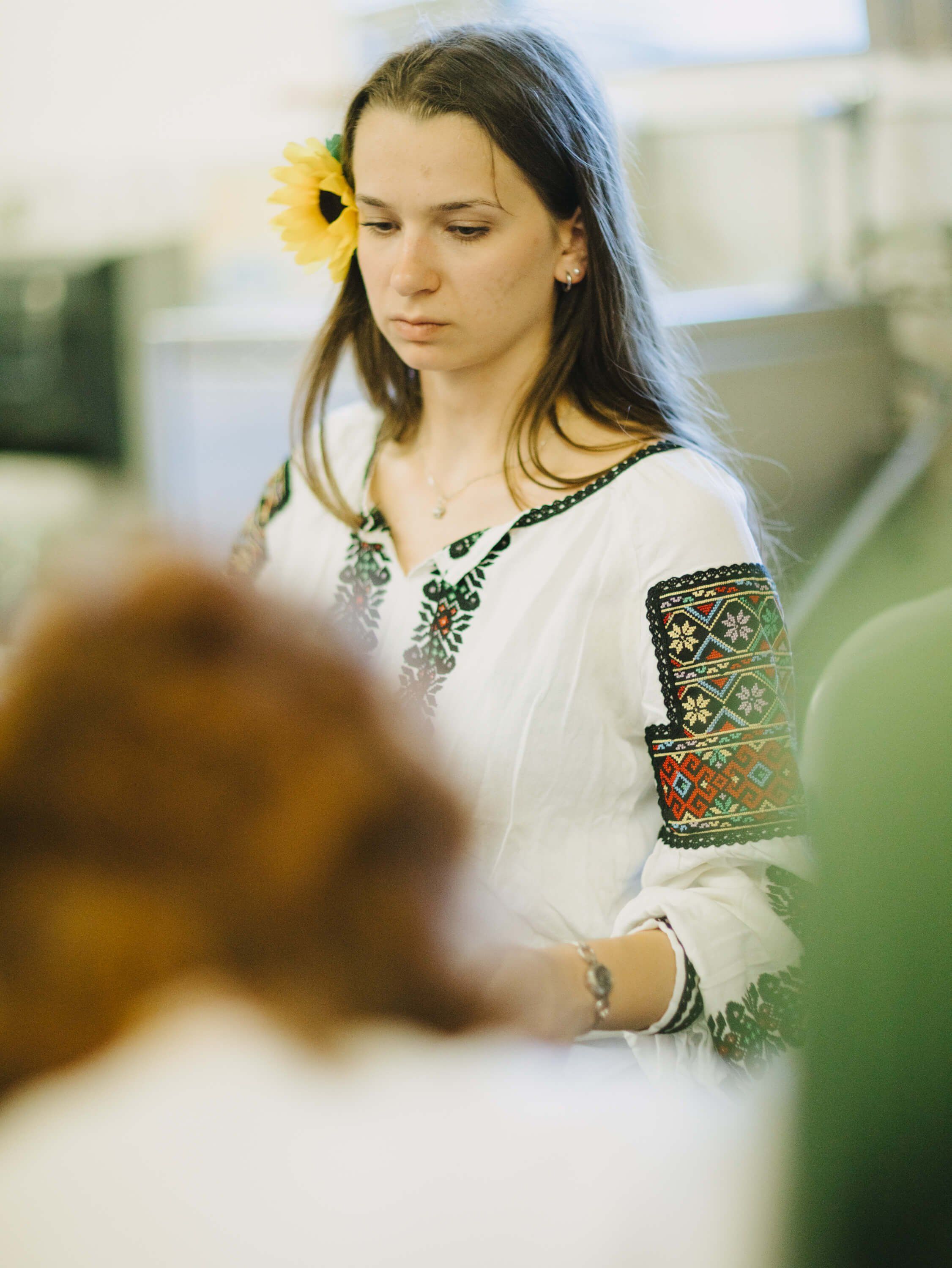 Woman in white embroidered Ukrainian shirt with yellow flower in hair, looking down thoughtfully at a blurred foreground object