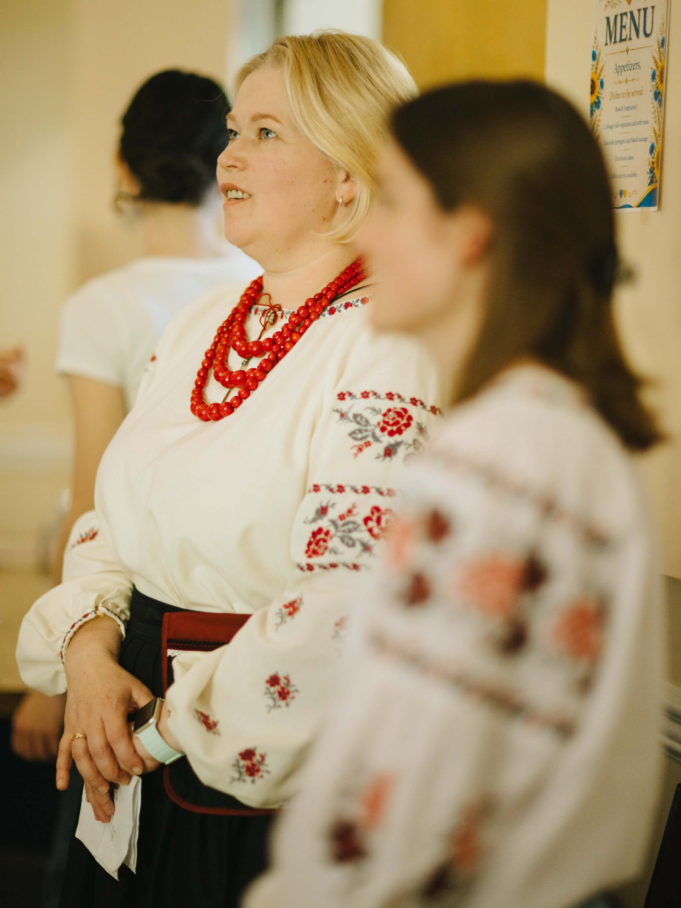 Woman in embroidered white blouse and red beaded necklace smiles while conversing at an indoor fundraiser event