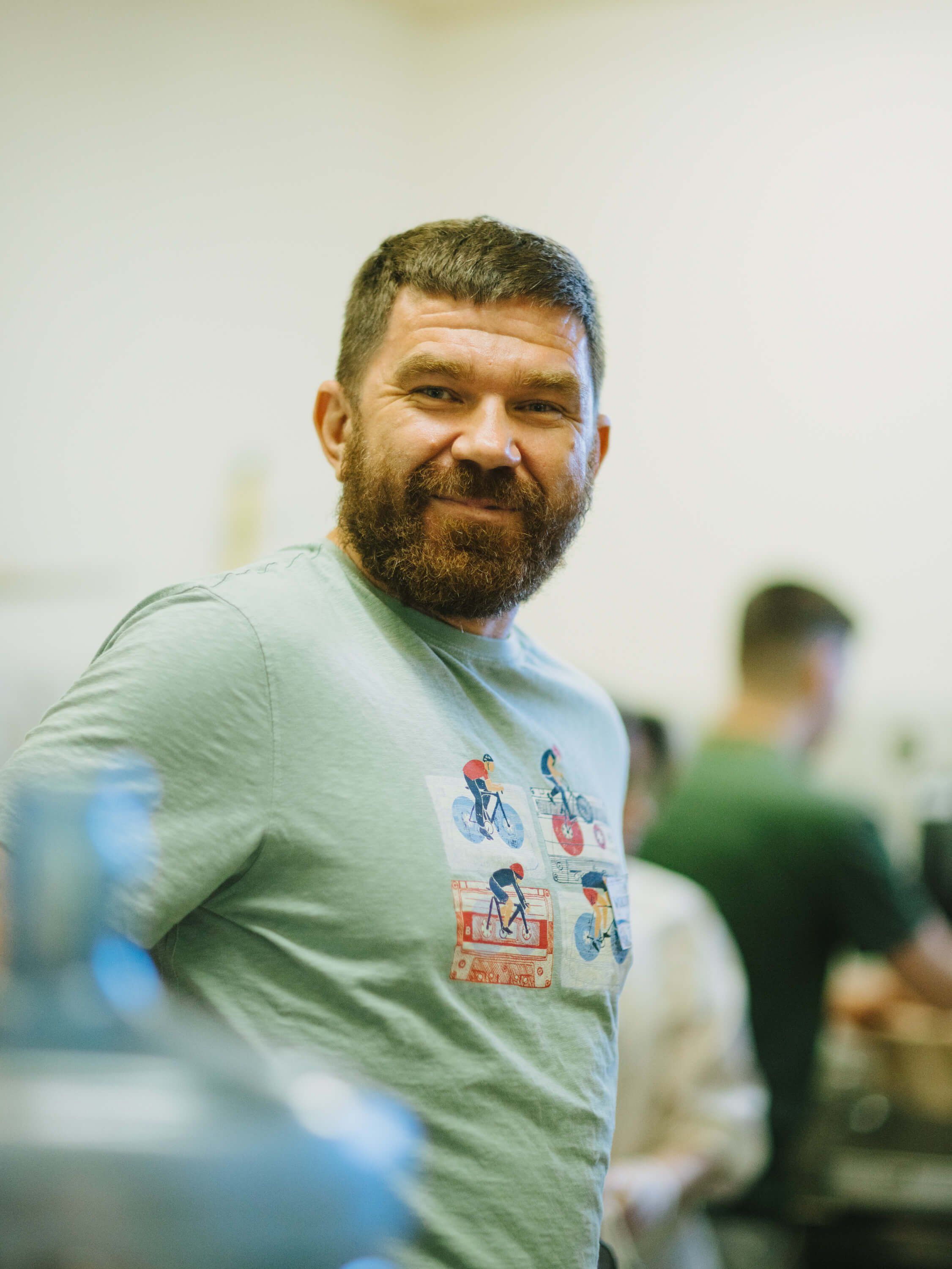 Bearded man in light green graphic tee smiles at camera during indoor fundraiser event with blurred attendees in background
