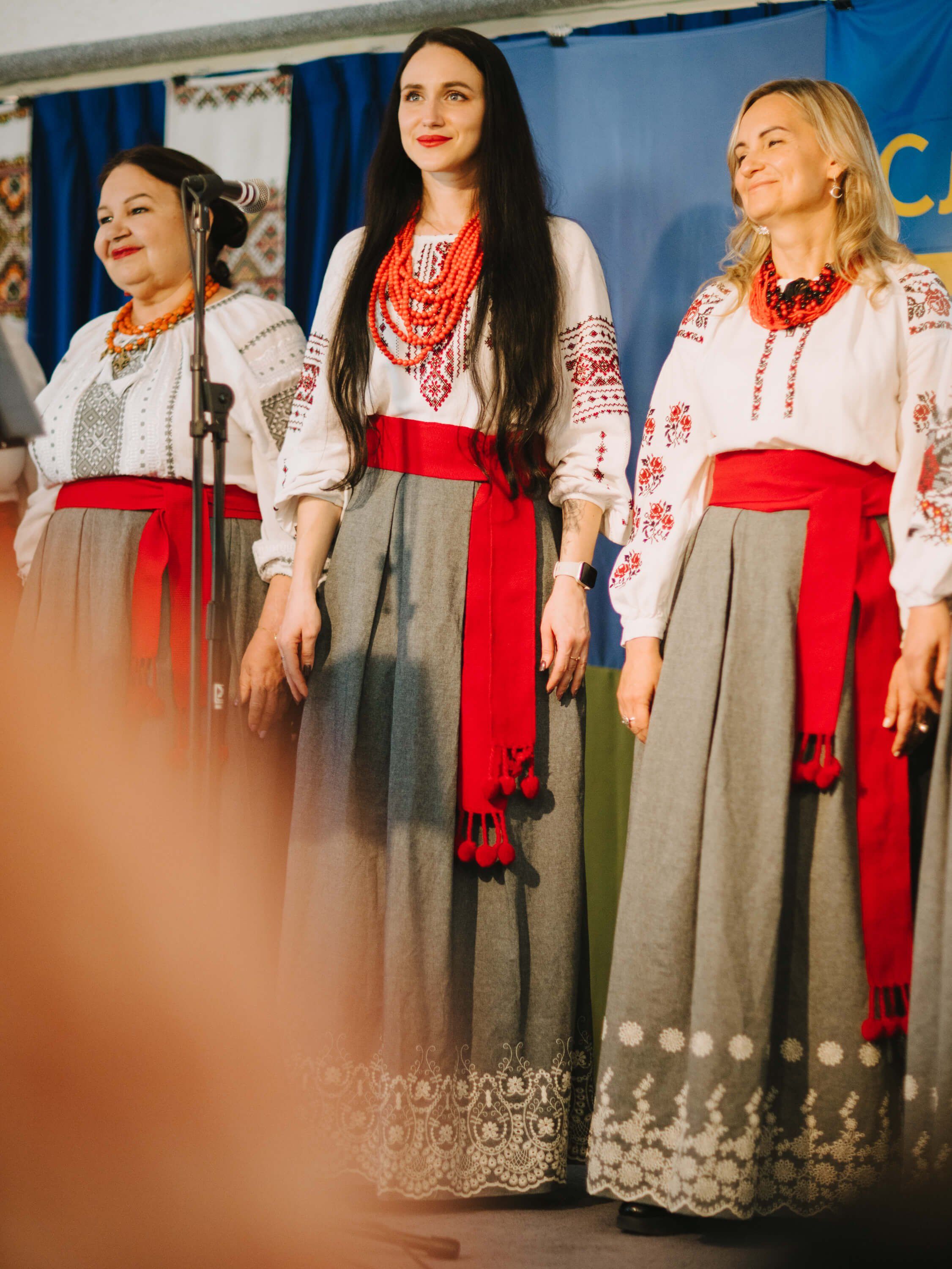 Three women in traditional Ukrainian embroidered white blouses and red-belted skirts stand together on stage at the fundraiser dinner