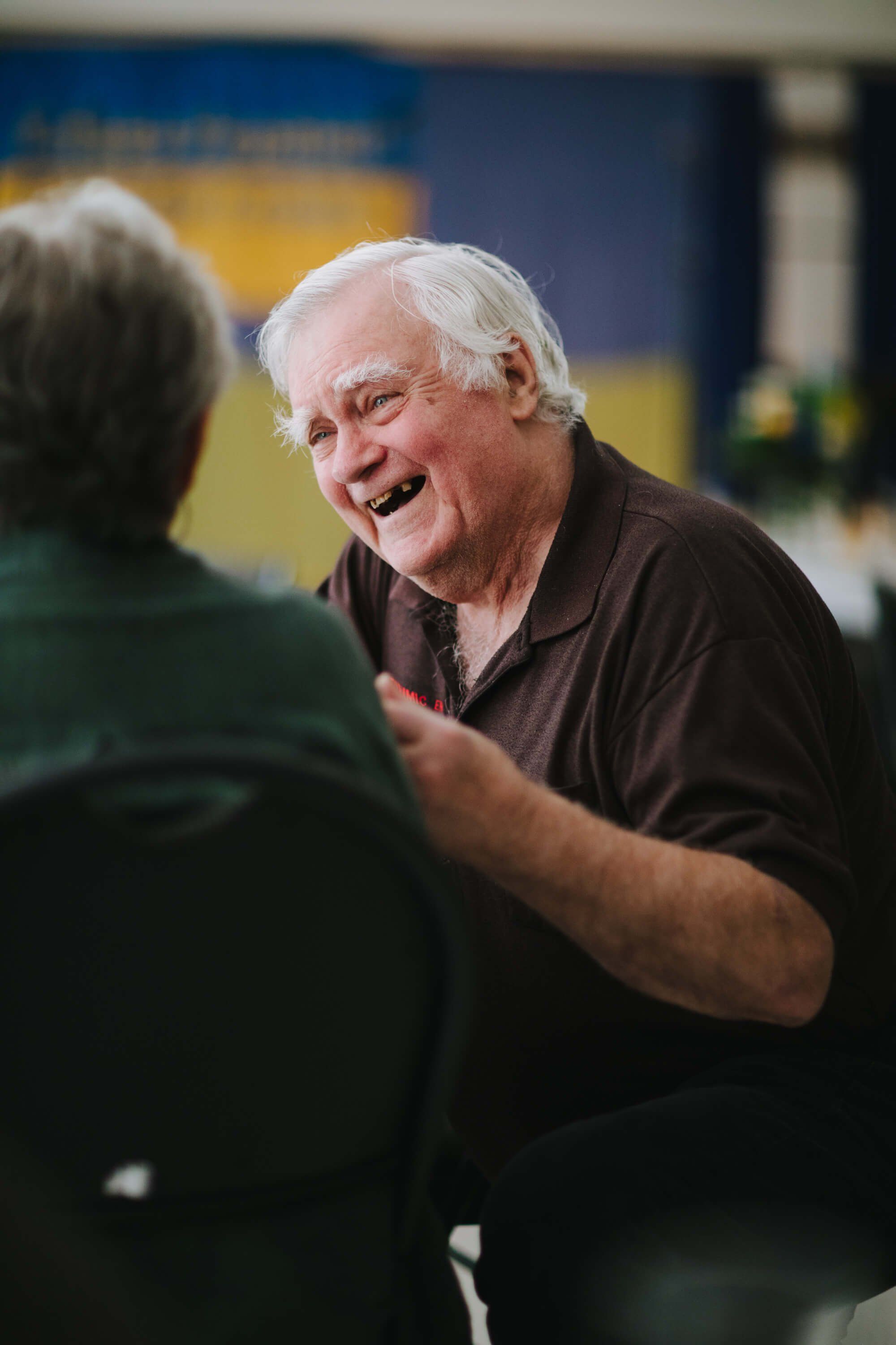 Older man with white hair in dark polo shirt laughs warmly while conversing with someone at an indoor event, blue and yellow backdrop visible behind