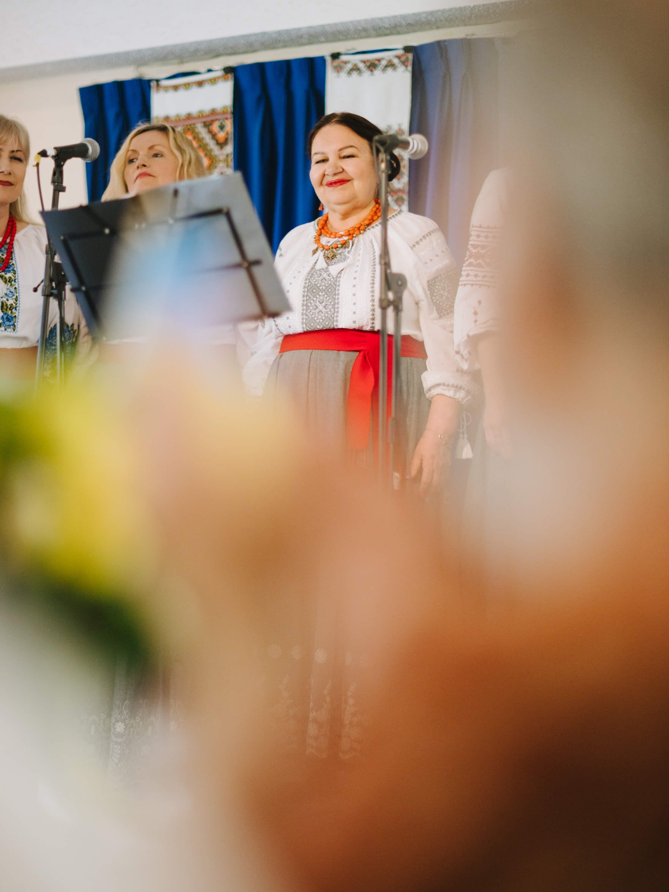 Woman in white embroidered shirt and red skirt smiles at microphone during fundraiser dinner, audience blurred in foreground