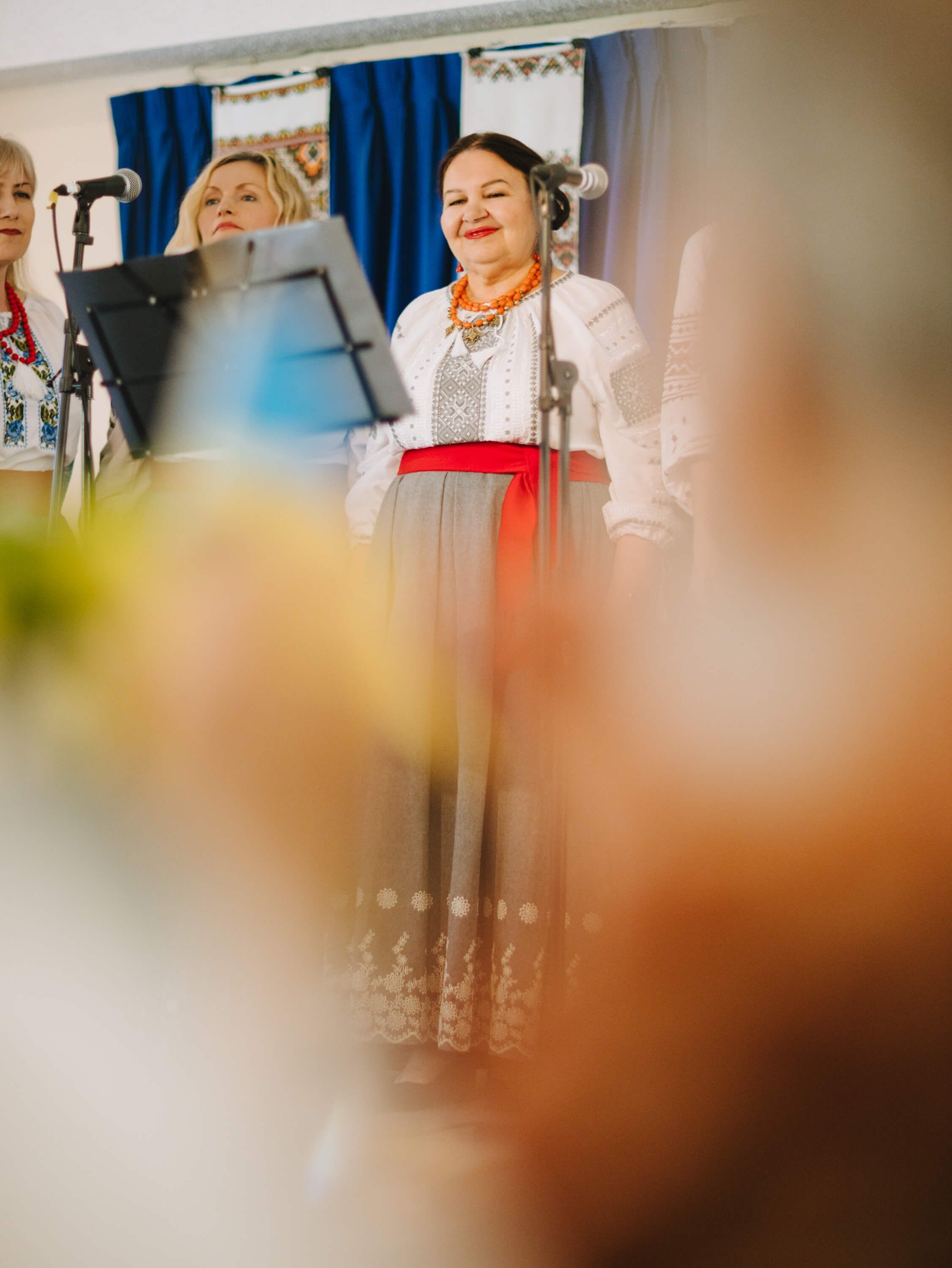 Woman in white embroidered dress with red belt speaks at microphone during Ukrainian fundraiser dinner event