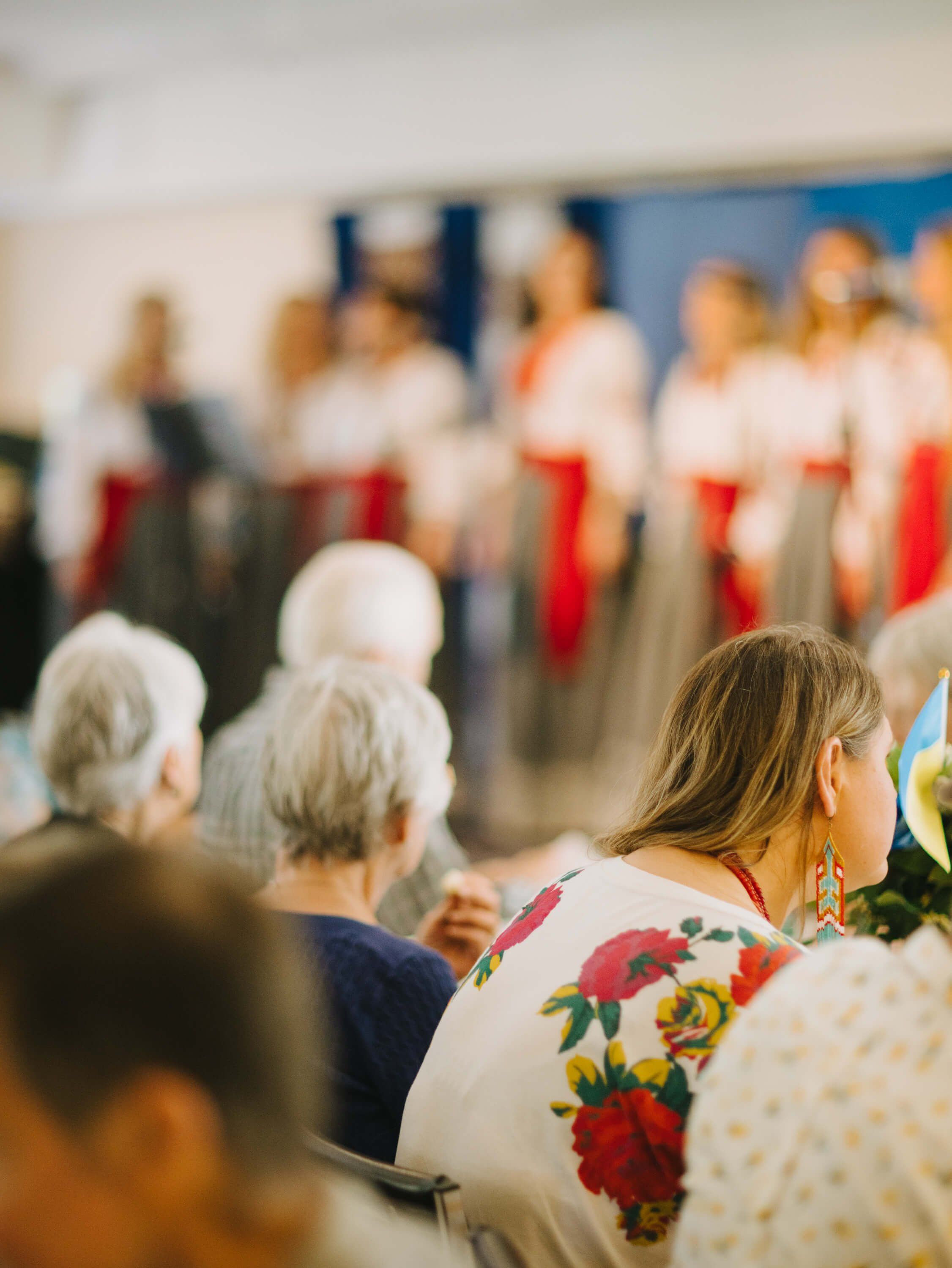 Attendees seated in foreground wearing floral patterns, facing a blurred choir in red and white robes performing on stage