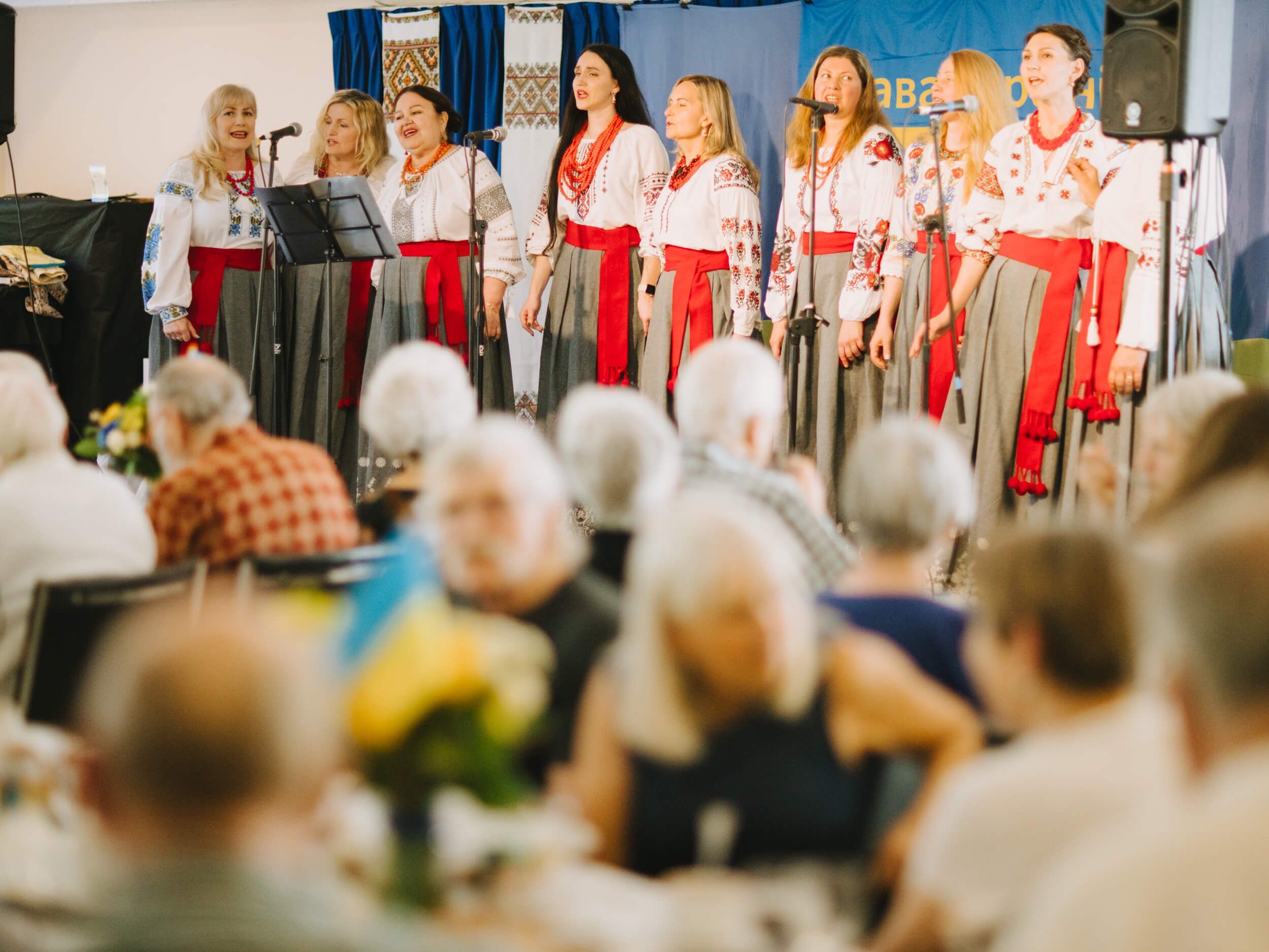 Group of women in traditional Ukrainian embroidered shirts and red skirts perform on stage before an audience of seated guests